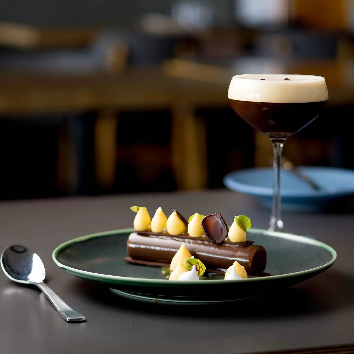 Elegant chocolate dessert on a green plate with cream, chocolate garnish, and edible flowers, accompanied by a dark cocktail with a creamy foam and chocolate pieces, on a dark table with a spoon nearby.