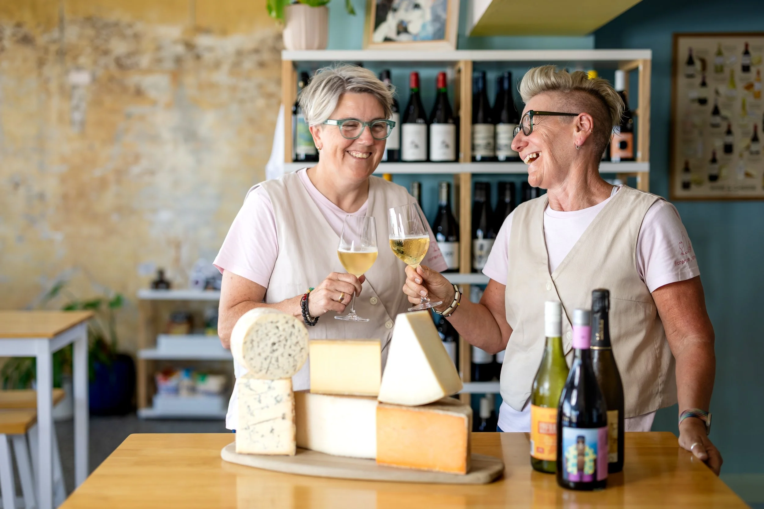 Two women smiling and holding wine glasses in a shop with cheese and wine bottles displayed on a counter.