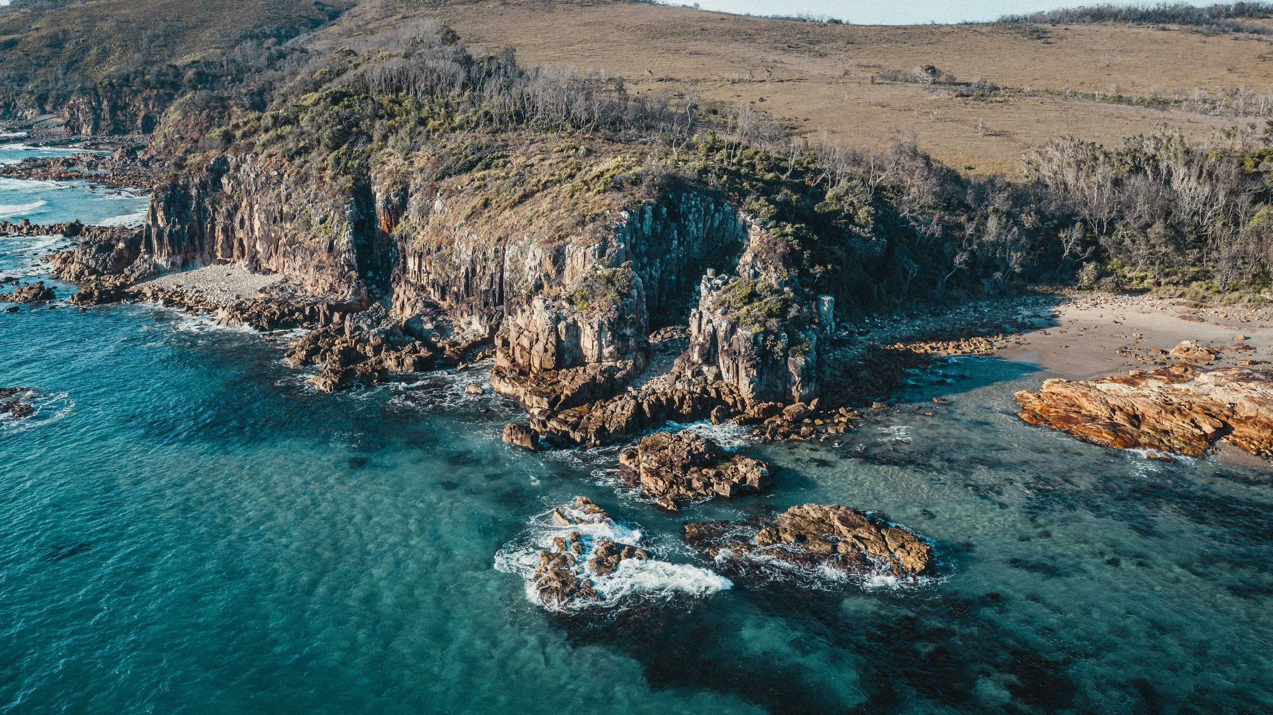 Aerial view of rocky coastline with cliffs, ocean waves, and sparse vegetation, with a sandy beach in the distance.