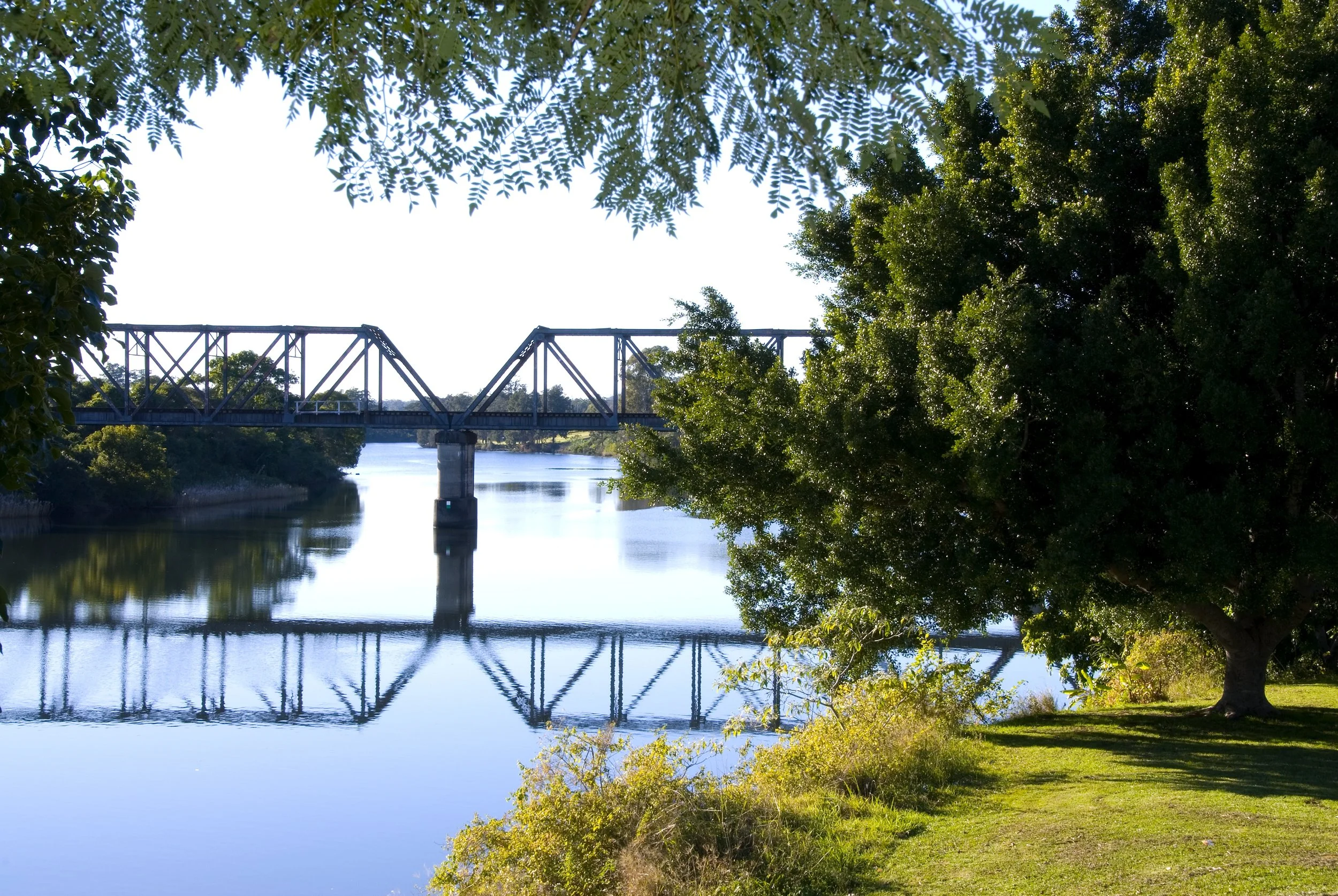 A metal train bridge spanning a calm river with lush green trees on both sides, under a partly cloudy sky.