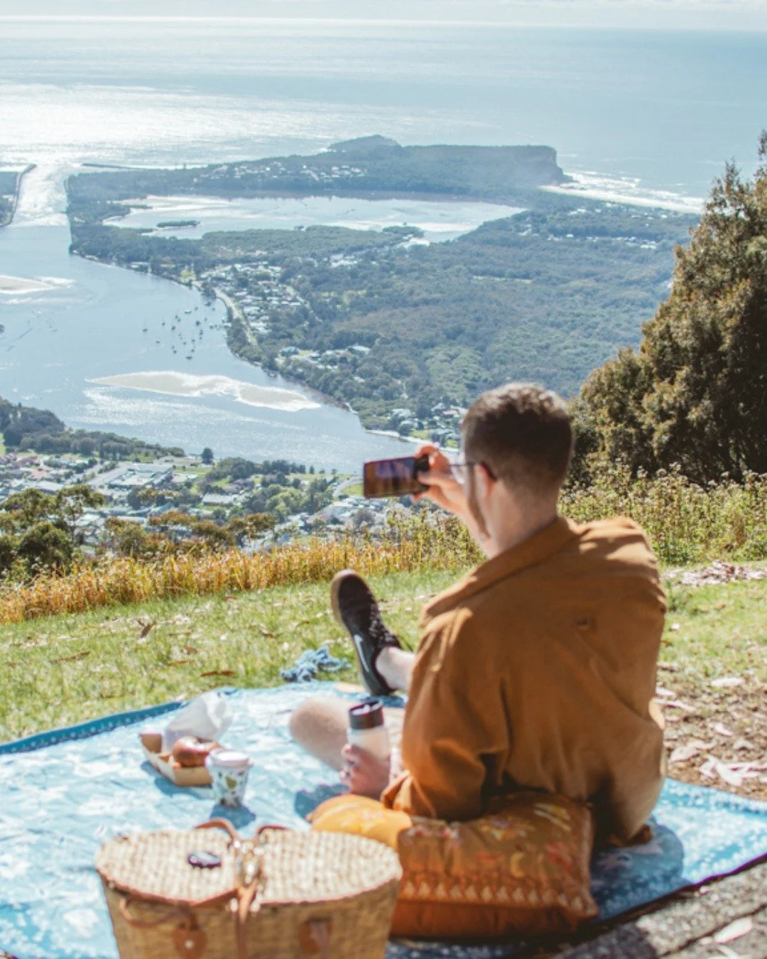 A person sitting on a blanket having a picnic on a grassy hill with a scenic view of a river, trees, and a distant coastline in the background.