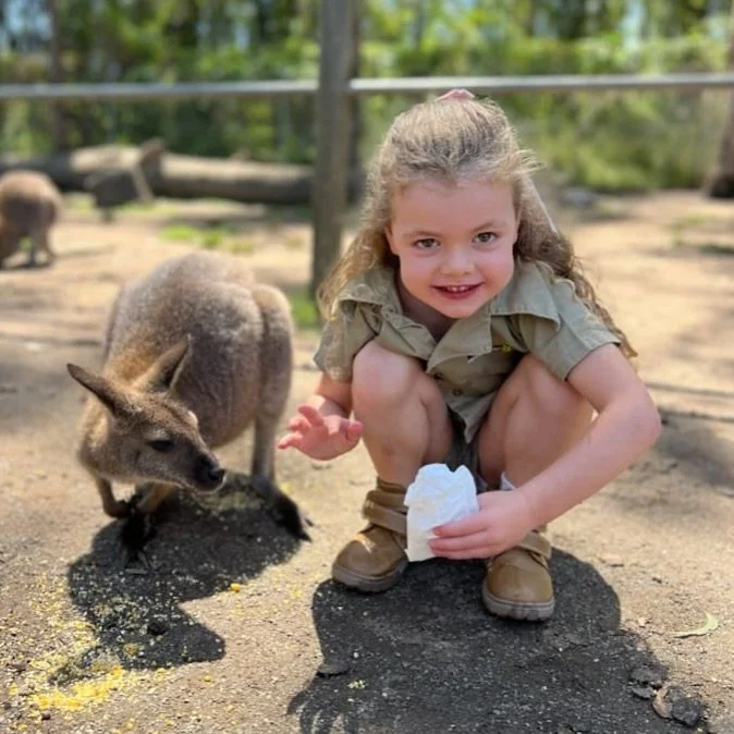 Young girl crouching on the ground next to a wallaby eating food at a zoo or wildlife sanctuary.