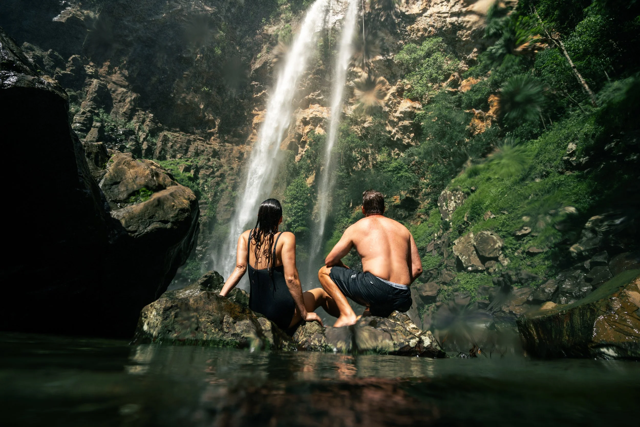 A man and woman sitting by a waterfall in a lush green forest, viewed from the water.