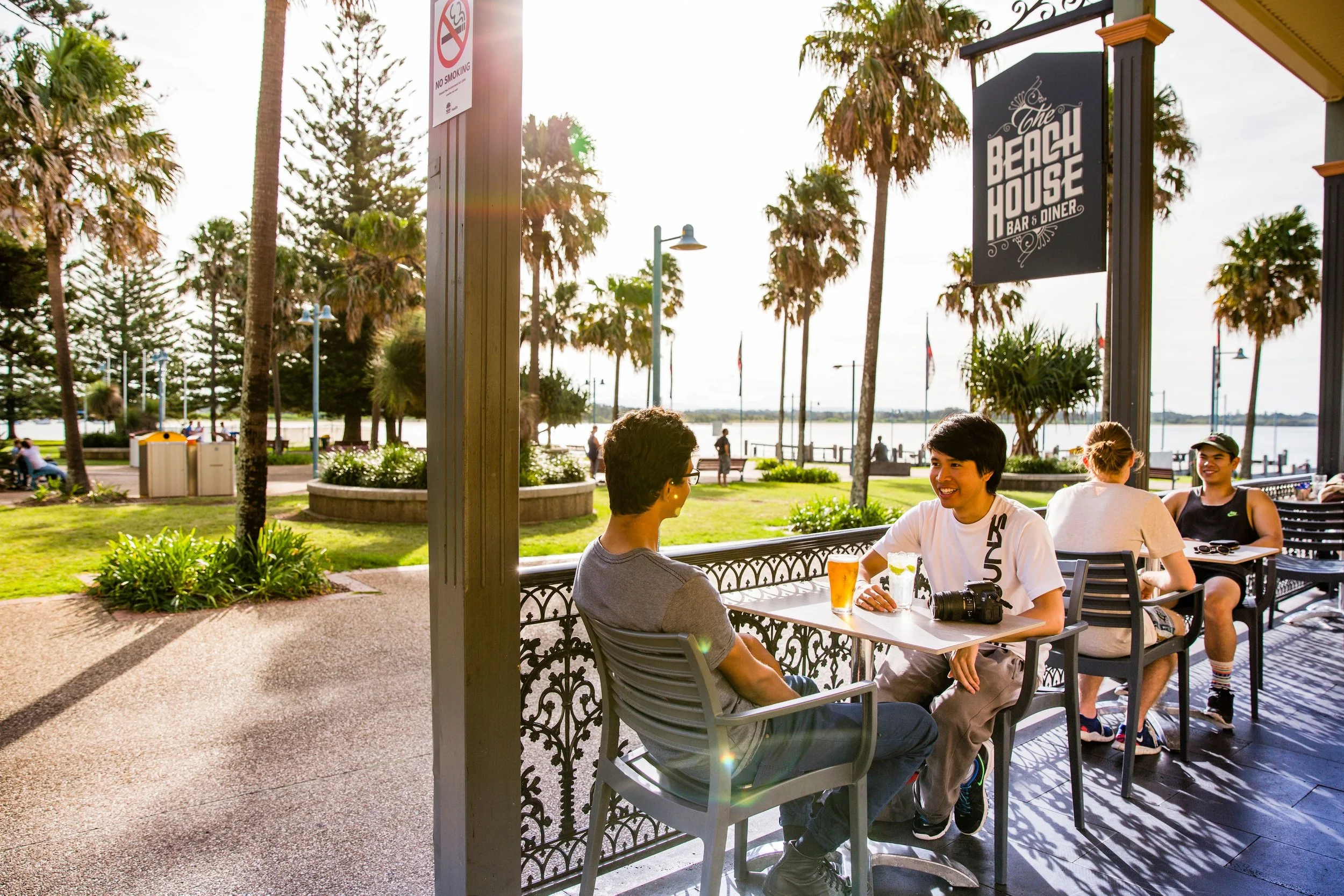People sitting at an outdoor patio by the water, with palm trees, a sign for The Beach House Bar & Diner, and a No Smoking sign in the background.