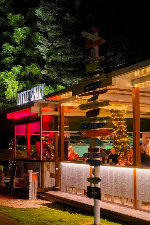 Night view of a modern outdoor dining area illuminated with warm lights, with a wooden signpost featuring colorful directional arrows, surrounded by trees and a lit building with a neon sign.