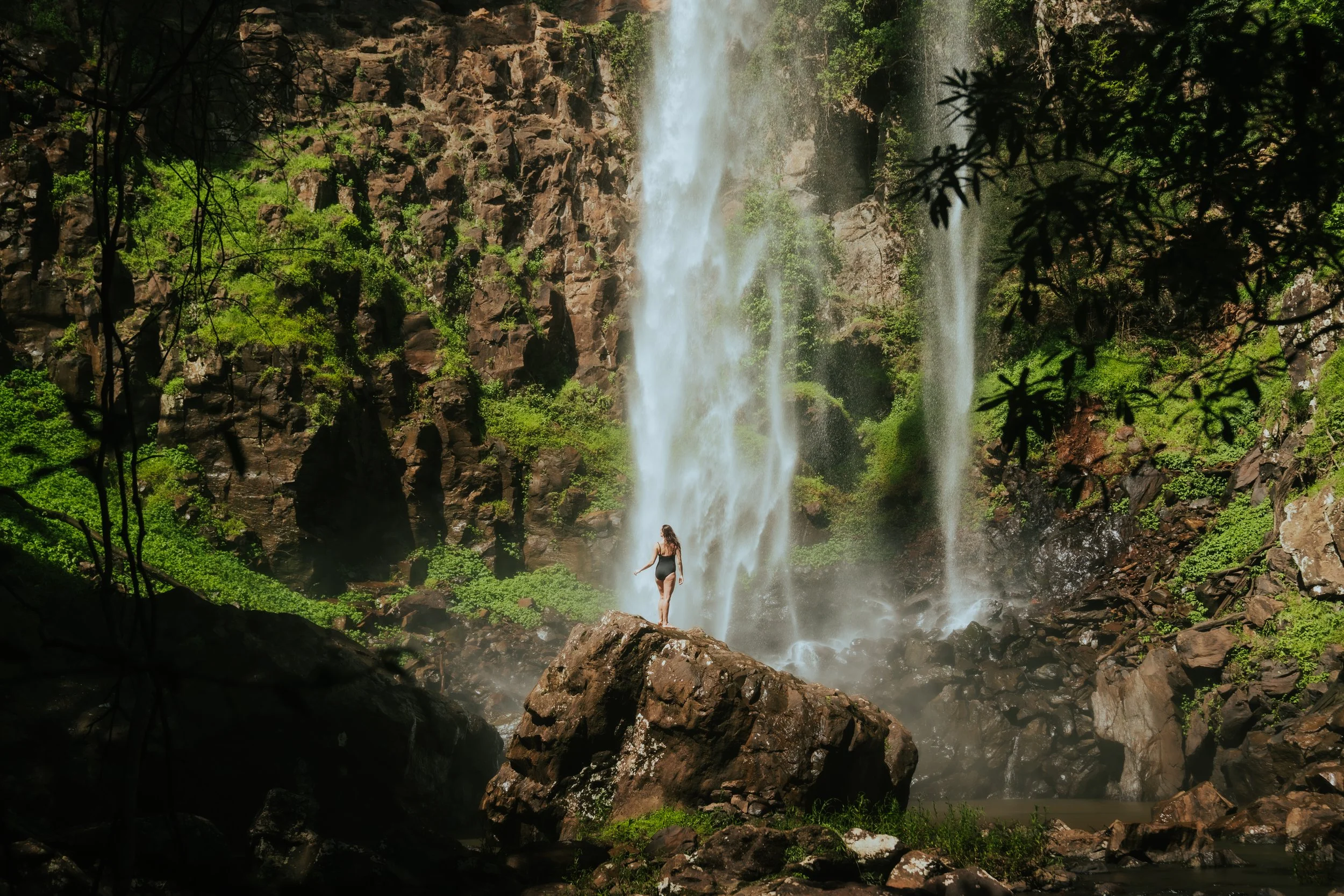 A woman in a swimsuit standing on rocks near a waterfall surrounded by greenery and trees.