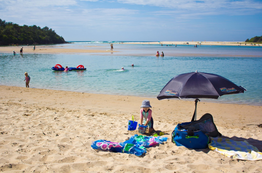 Child playing on sandy beach under a large umbrella with water, swimmers, and boats in background.