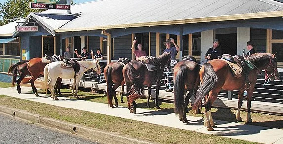 Several horses tied to a post in front of a building with people standing and riding horses.