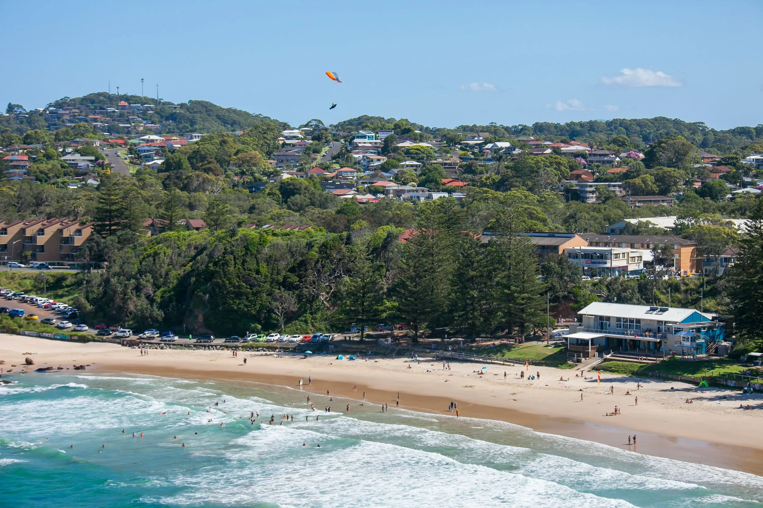 A coastal scene featuring a sandy beach with people swimming and relaxing, ocean waves, and a hillside covered with residential houses and trees, dotted with colorful kites flying in a blue sky.