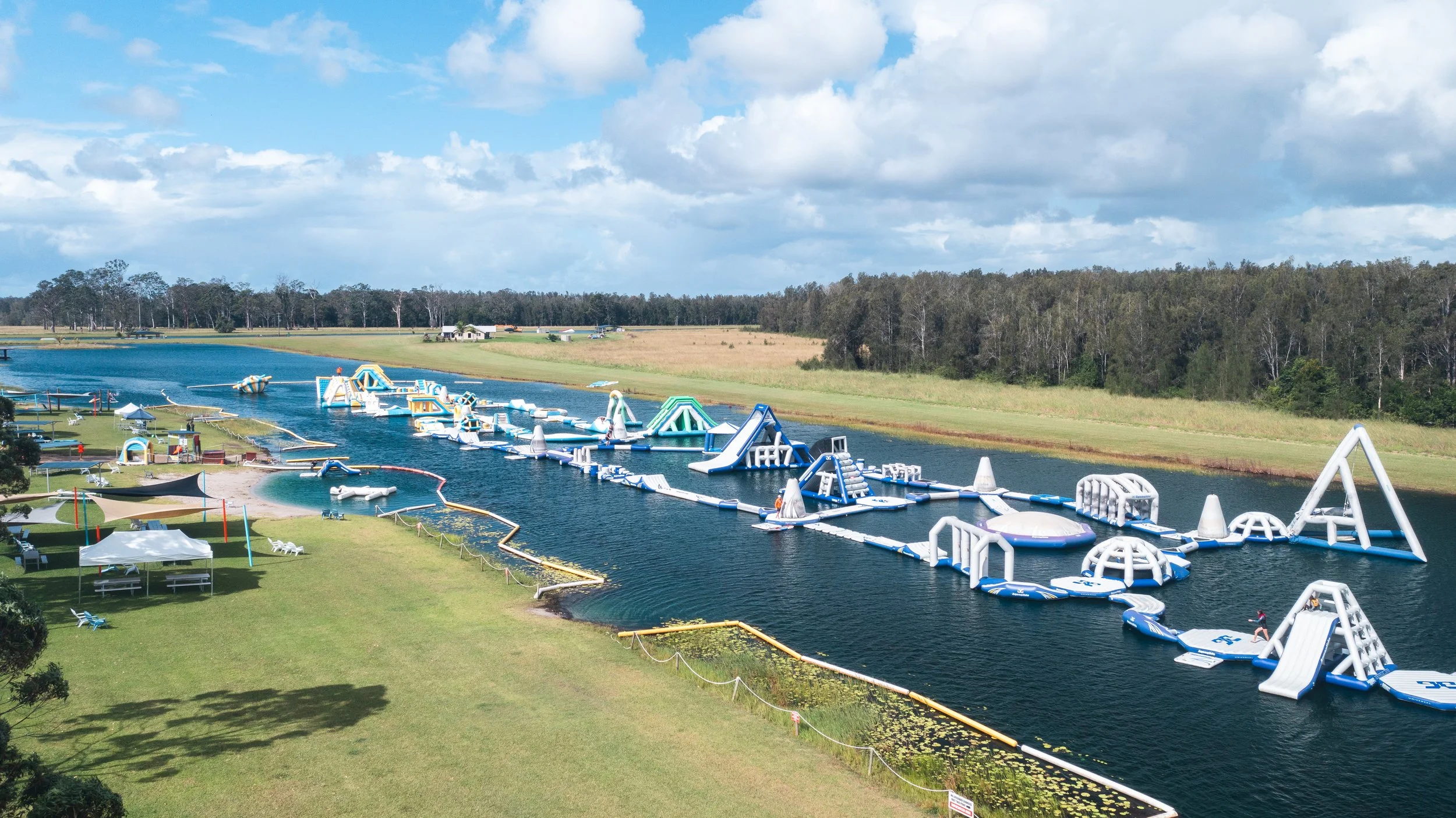 Large outdoor water park with inflatable floating obstacle courses on a river, with grassy area and trees in the background, under a partly cloudy sky.