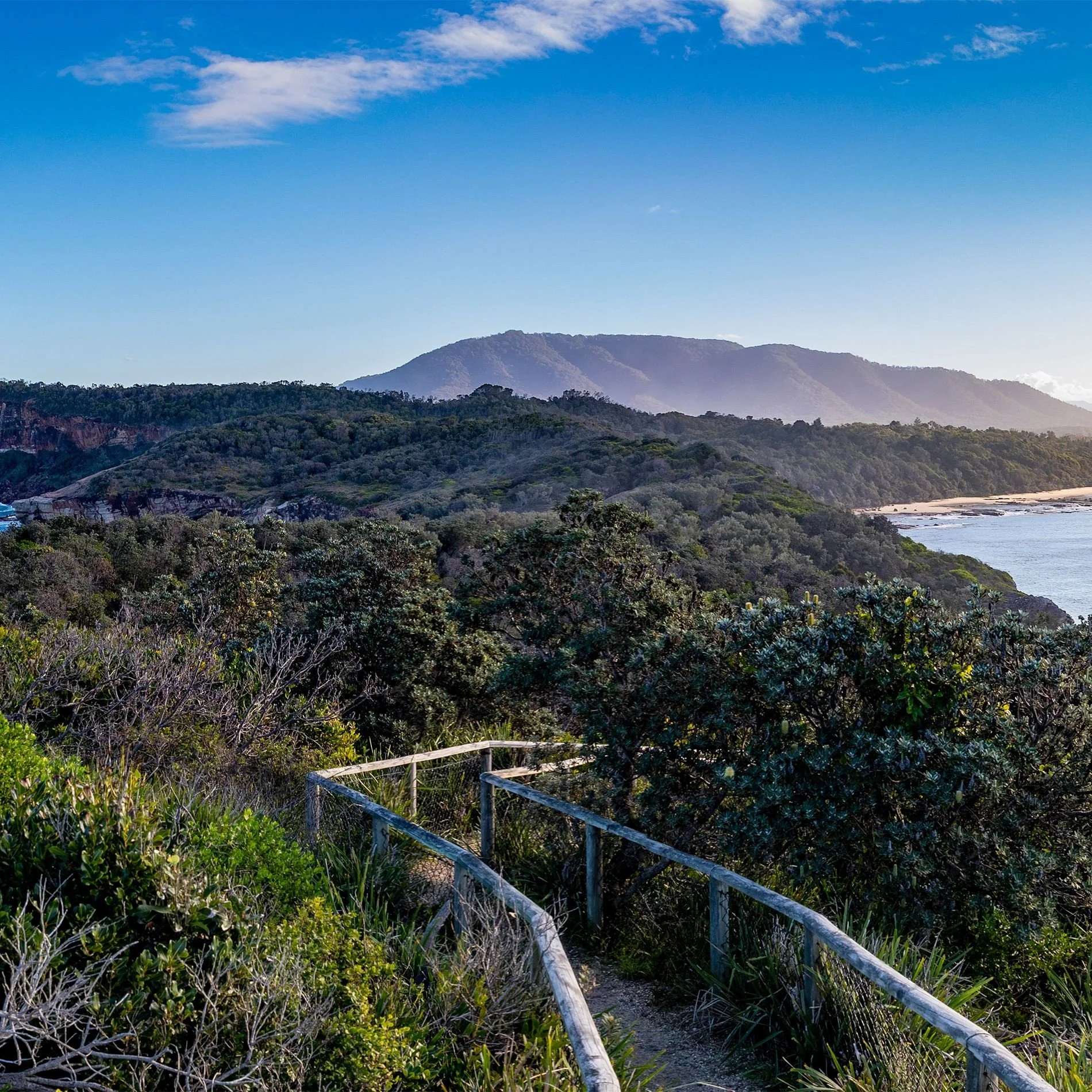 A coastal landscape with hills covered in green vegetation, a sandy beach, and blue ocean water under a clear sky with some clouds.