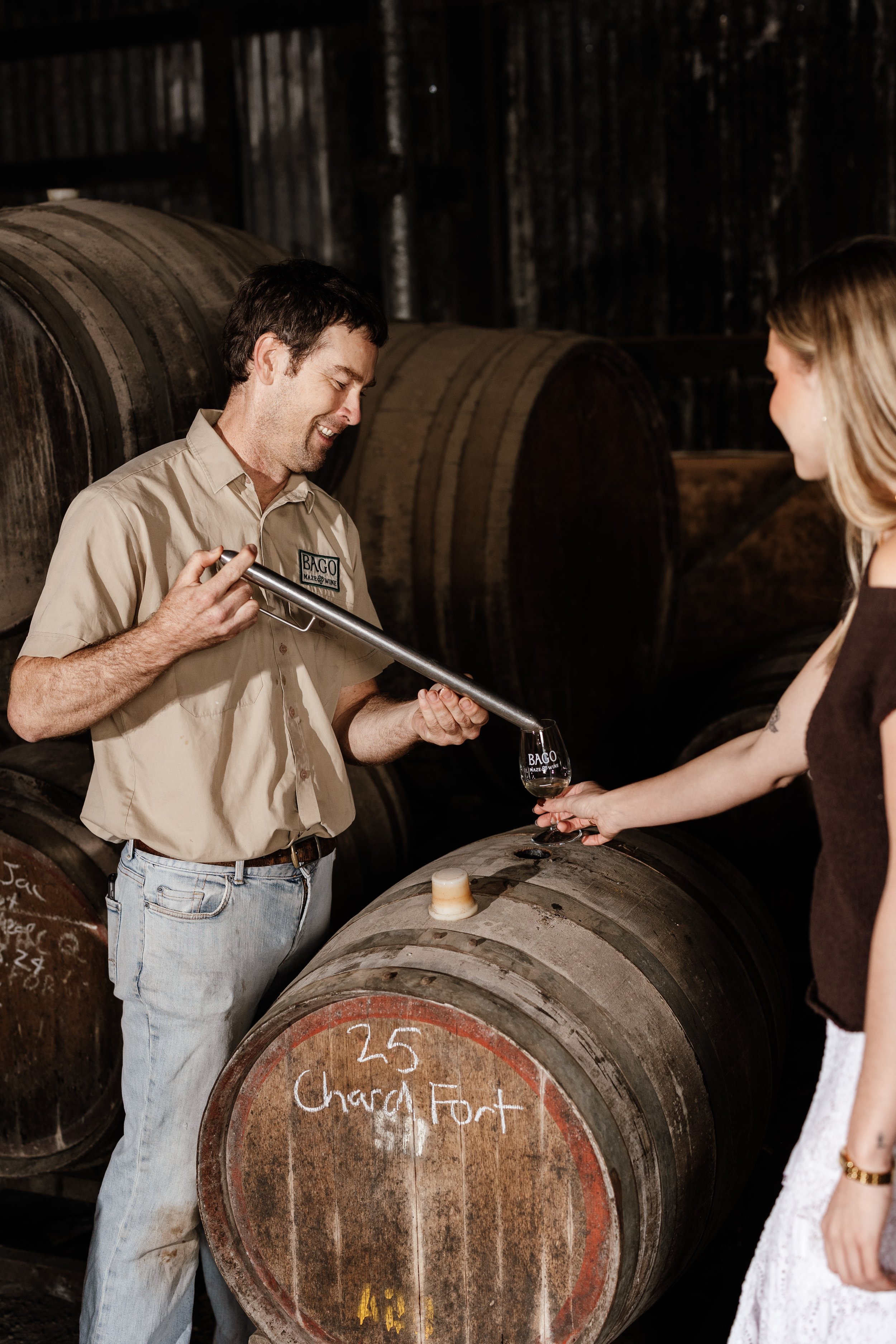 A man in a beige shirt and light blue jeans is pouring wine from a pipe into a woman's glass at a winery. They are standing next to wine barrels, with one barrel labeled '25 Charca Fort.'