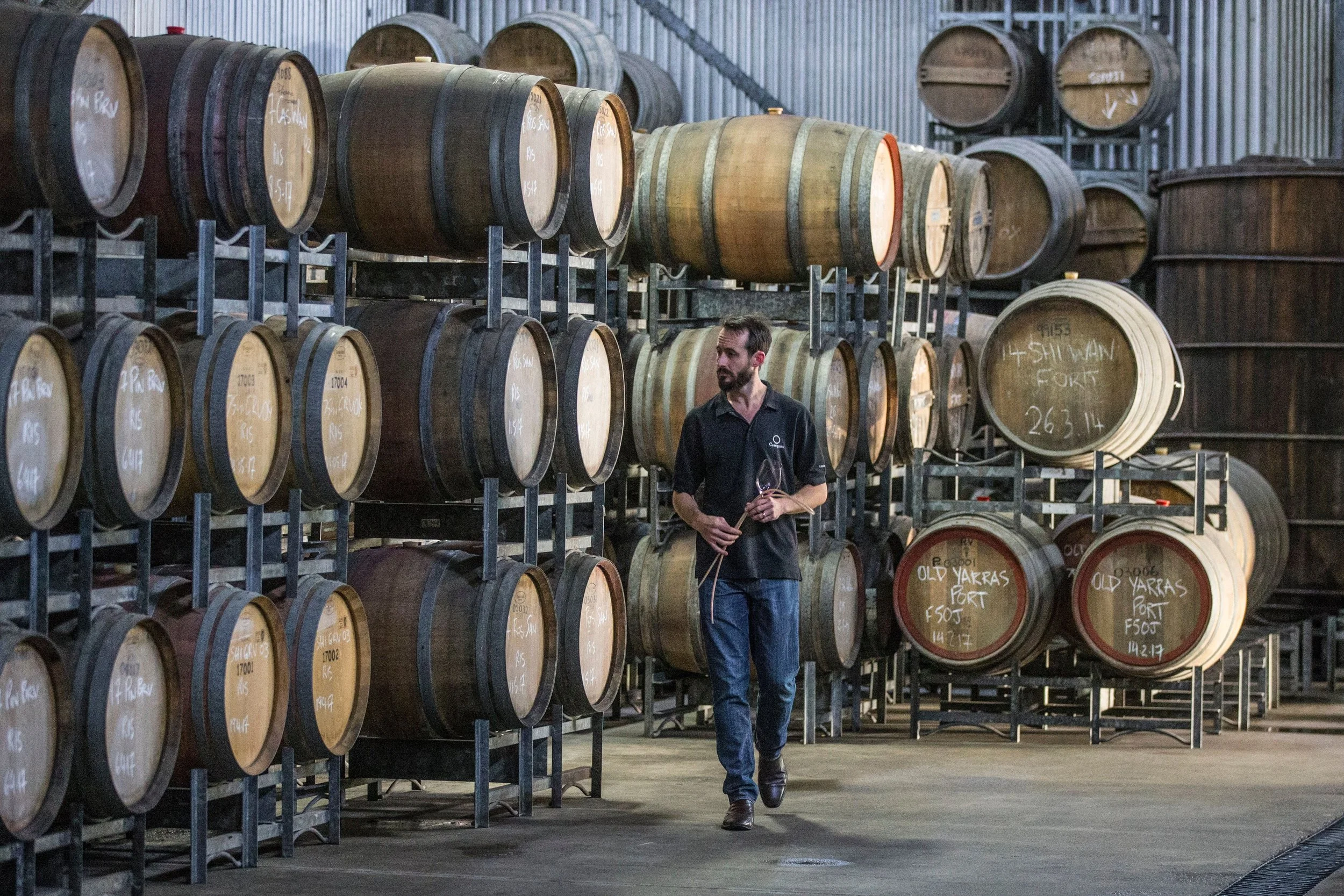 A man with a beard and dark hair walking through a warehouse filled with stacked wine barrels on metal racks. The barrels have handwritten labels.