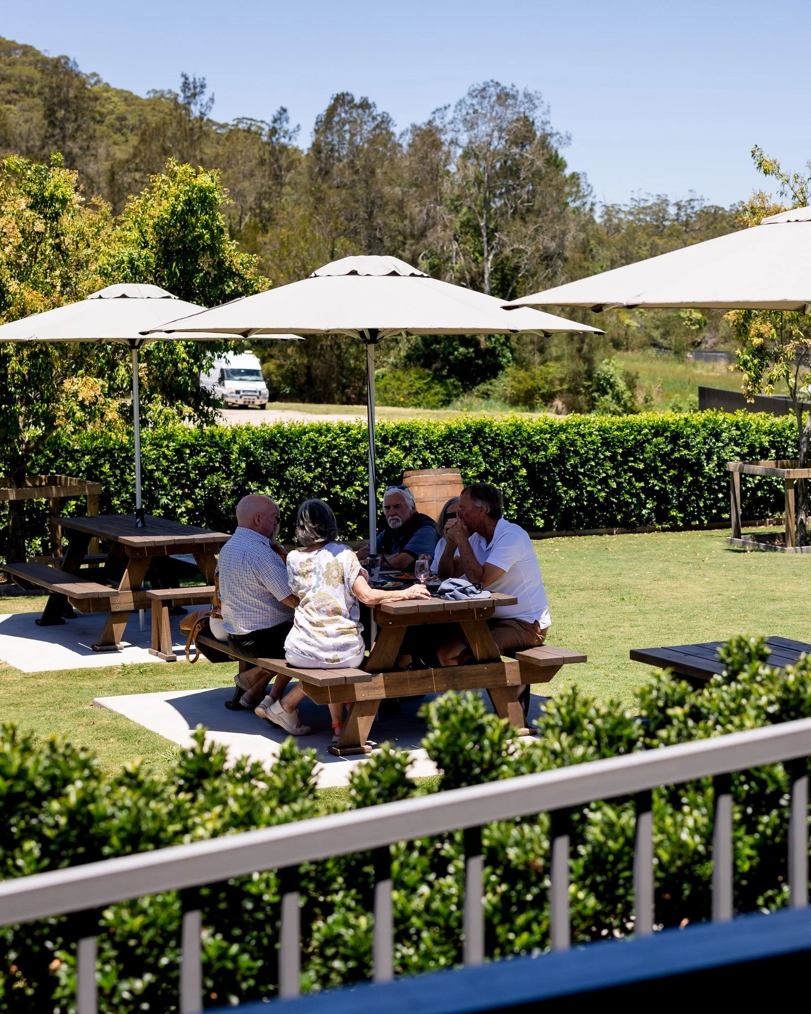 People sitting and talking at a picnic table beneath large white umbrellas in a green outdoor setting with trees and a hedge in the background.