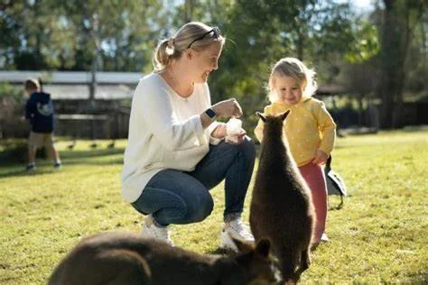 A woman and a young girl interacting with a small bear at a park during daytime, with other children in the background.