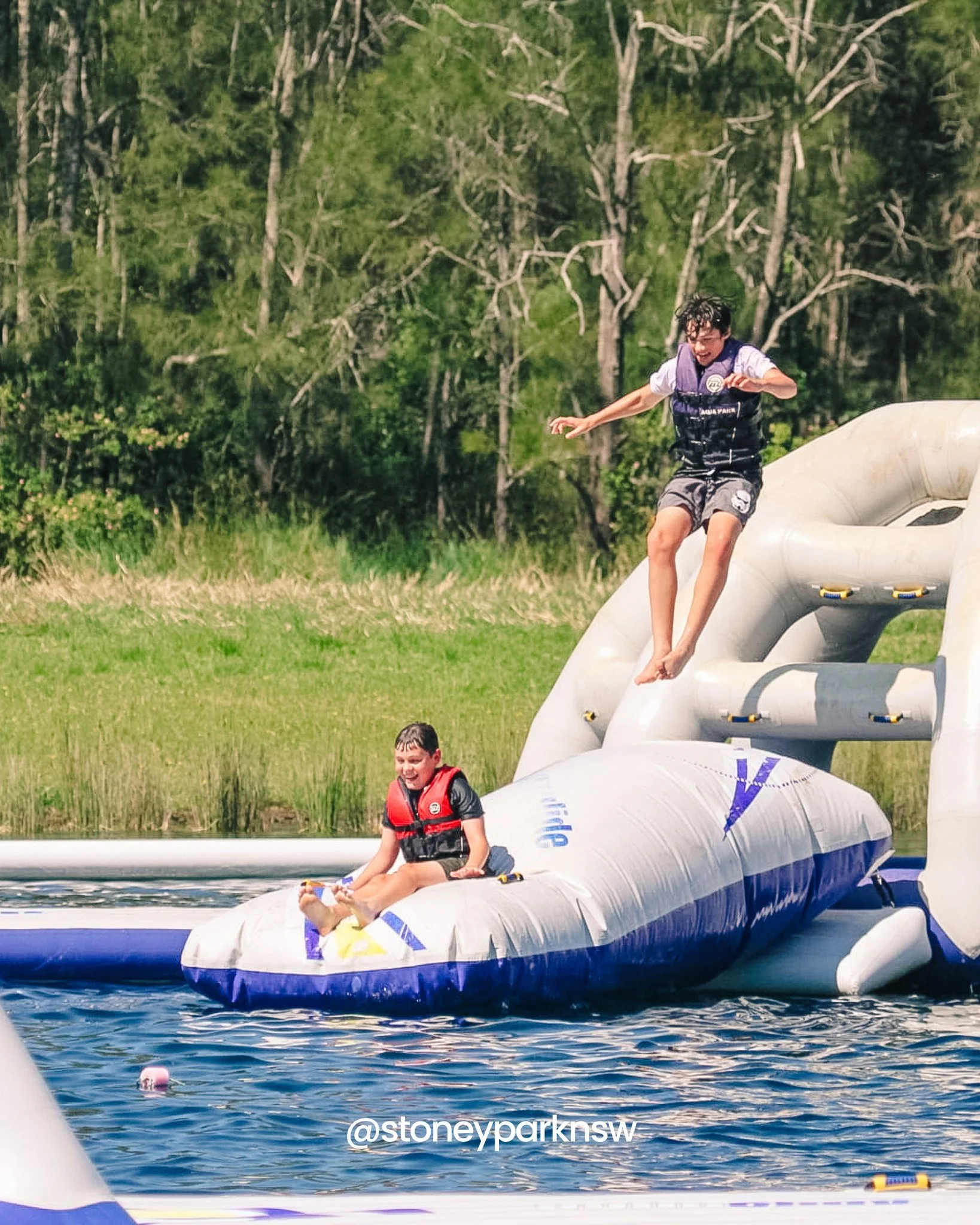 Two boys enjoying a ride on an inflatable boat on a body of water, with one boy jumping off the boat and another sitting on it, surrounded by greenery and trees.