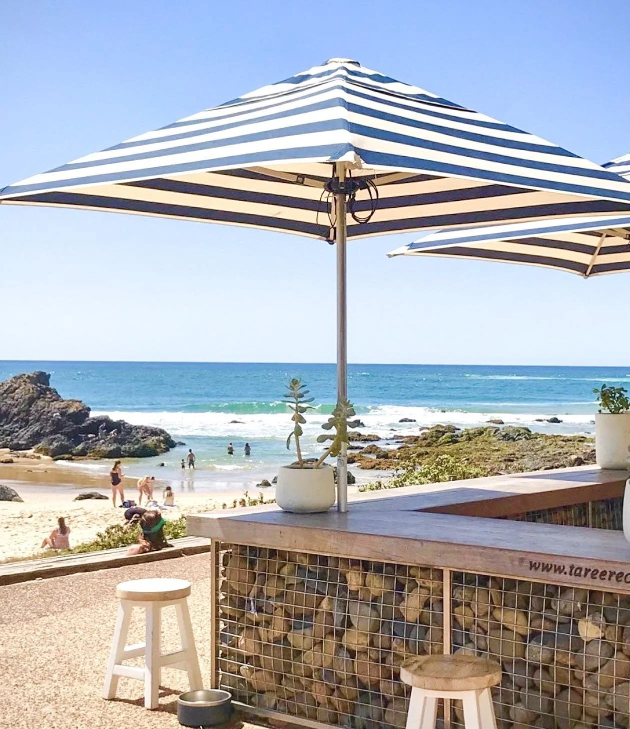 Beach scene with striped blue and white umbrellas, people swimming and relaxing near rocks, ocean waves, clear blue sky, and potted plants on a wooden deck.
