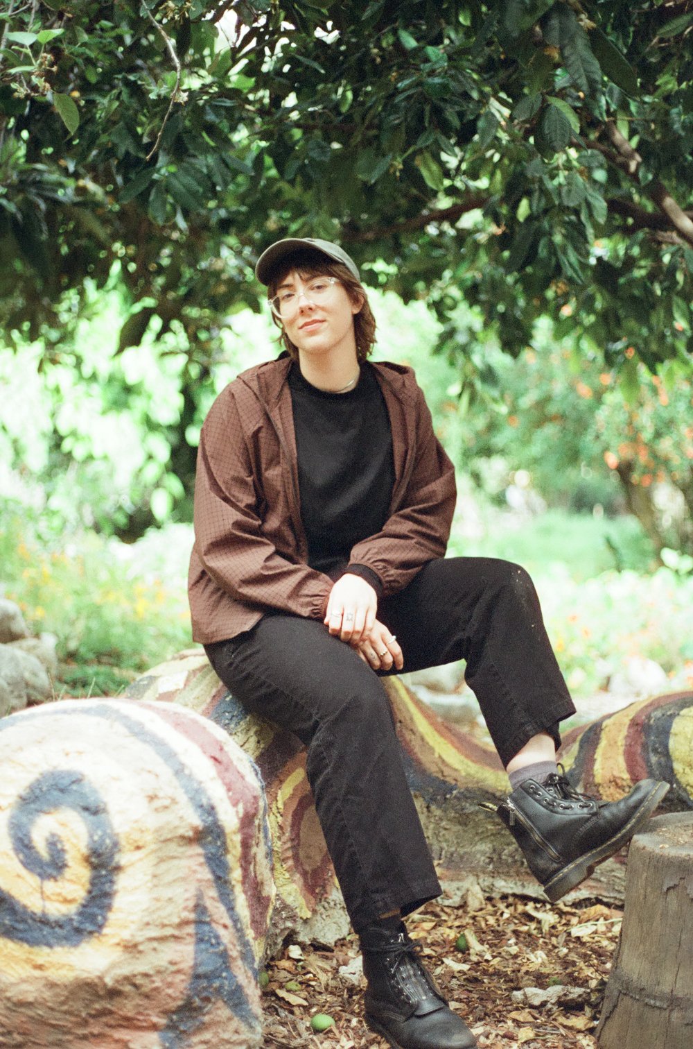 A person with short hair, wearing glasses, a black shirt, a brown jacket, black pants, and black boots, sitting on a painted stone in a garden surrounded by greenery and trees.