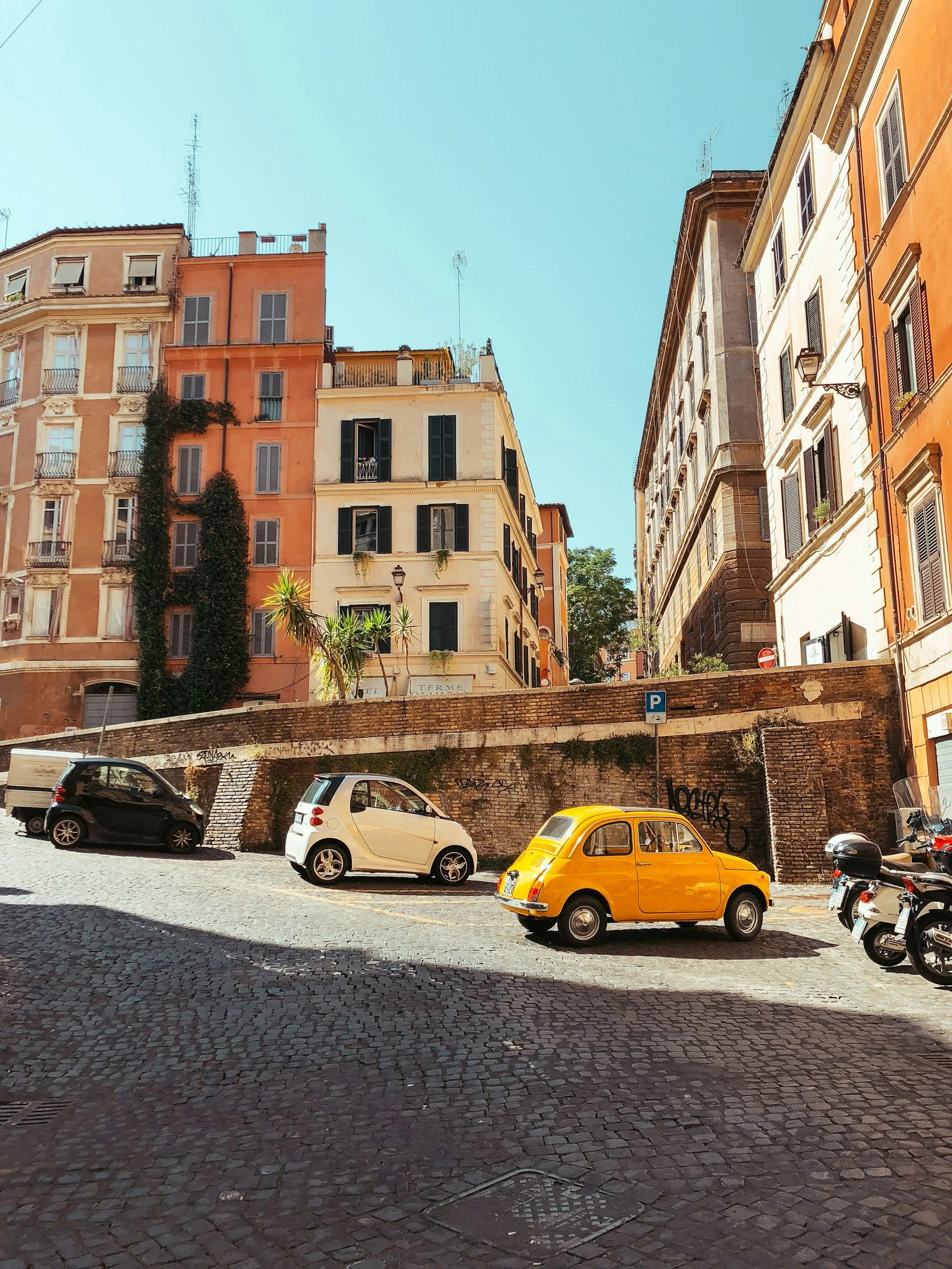 Colorful European city street with cobblestone pavement, parked cars including a yellow classic car, and multi-story buildings with shutters under a clear blue sky.