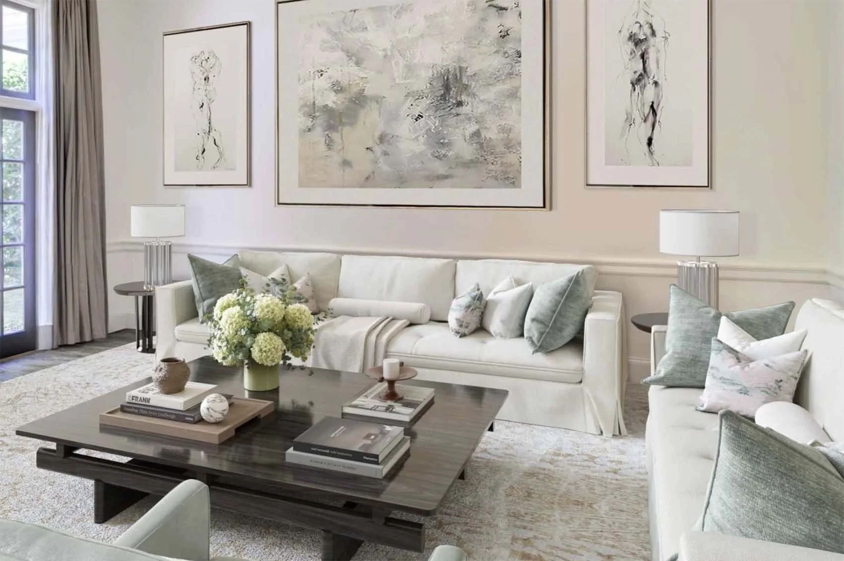 Living room with white sofas, decorative pillows, a dark wooden coffee table with books and a flower arrangement, side tables with lamps, large abstract paintings on the wall, and a glass door with curtains.