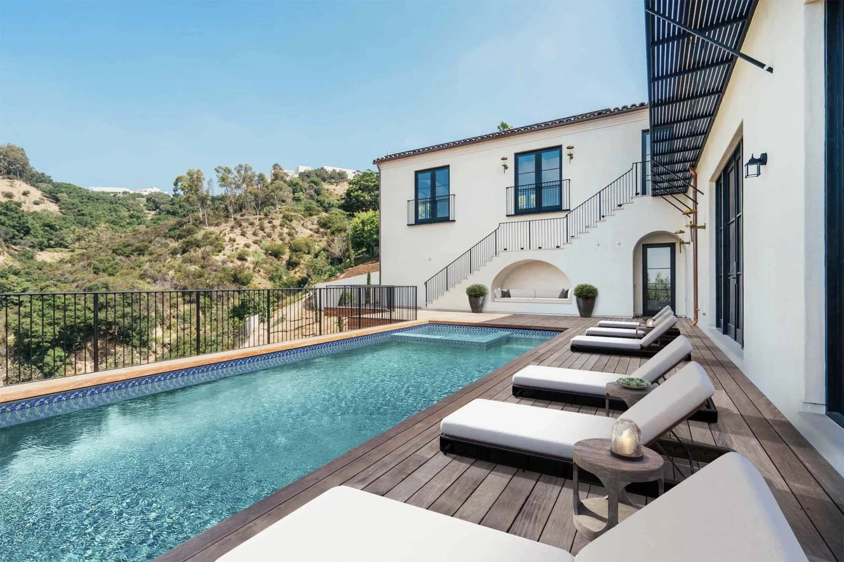 Outdoor swimming pool with lounge chairs on a wooden deck next to a modern white house with balcony doors, overlooking a hill with green trees and bushes under a blue sky.