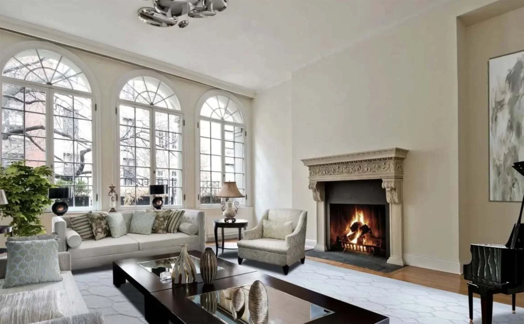 Living room with three large arched windows, a fireplace with a fire, a cream-colored sofa with multiple pillows, a small armchair, a wooden coffee table with decorative vases, a side table with a lamp, and a grand piano. The room has light walls, a white rug, and hardwood floors.
