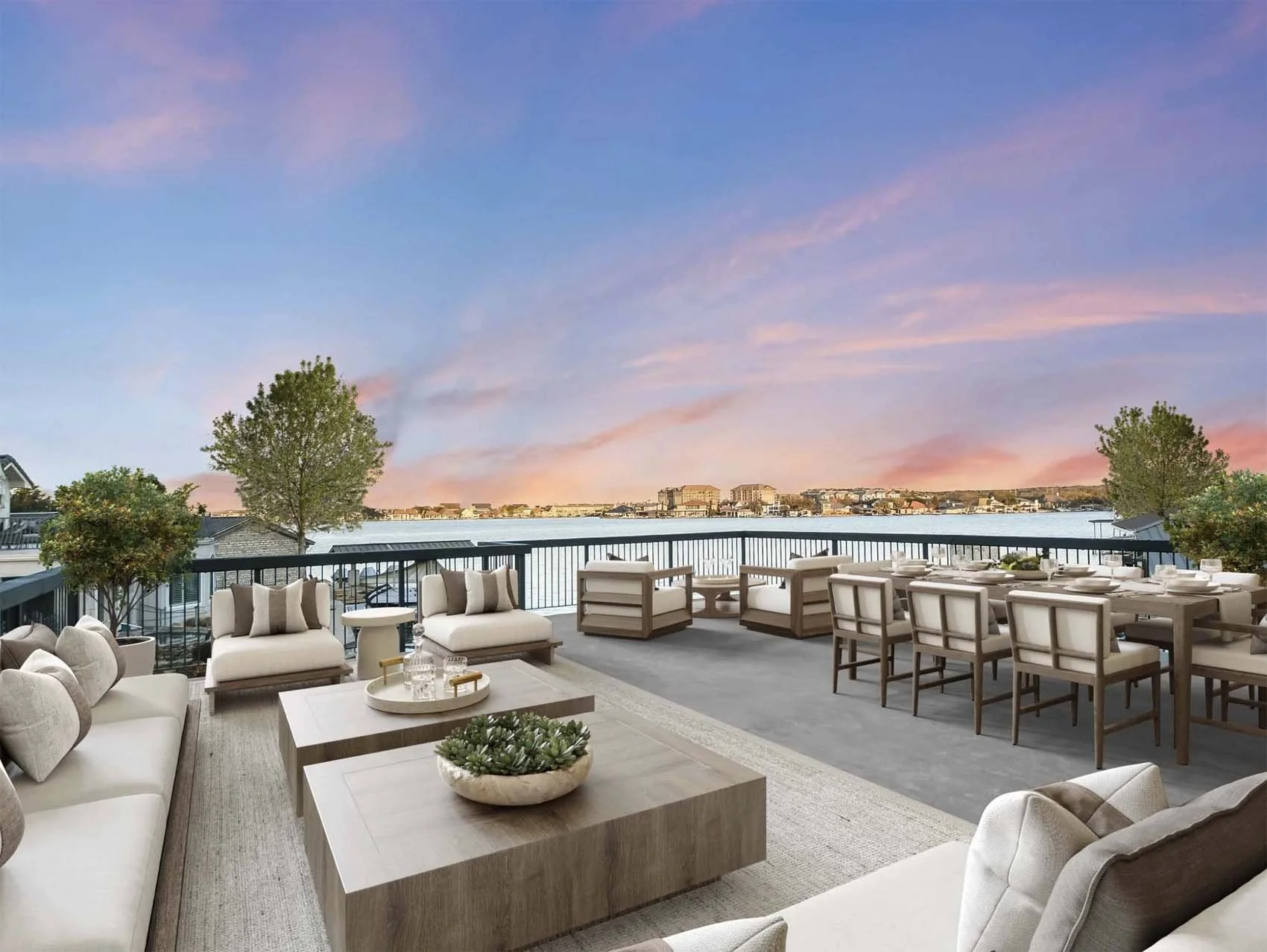 A spacious outdoor patio with beige cushioned seating, wooden tables, and a large dining table set for a meal. The patio is overlooking a body of water with buildings in the distance, under a colorful sky at sunset.