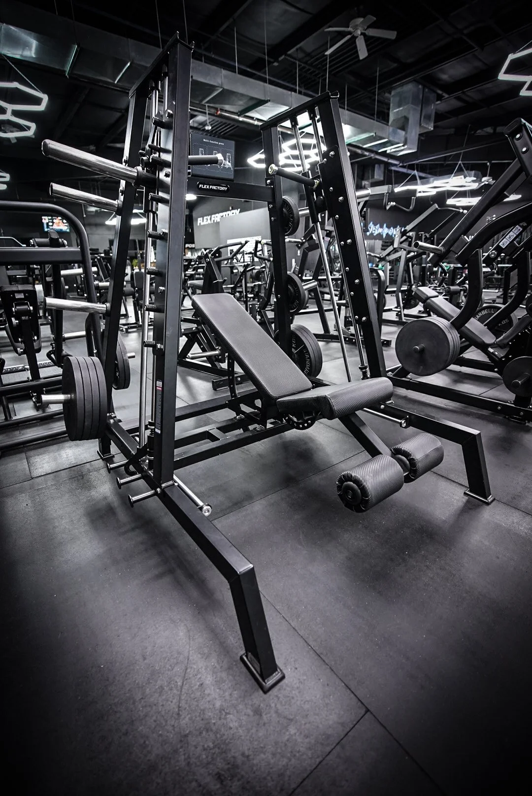 A black and gray gym equipment bench in a fitness center with various weight machines and equipment in the background.