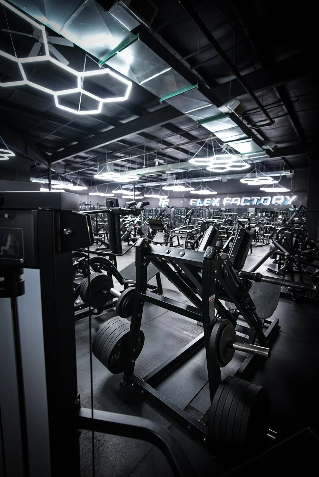 Interior of a modern gym with various exercise equipment and illuminated hexagon-shaped ceiling lights, featuring a sign that reads 'FLEX FACTORY' in the background.