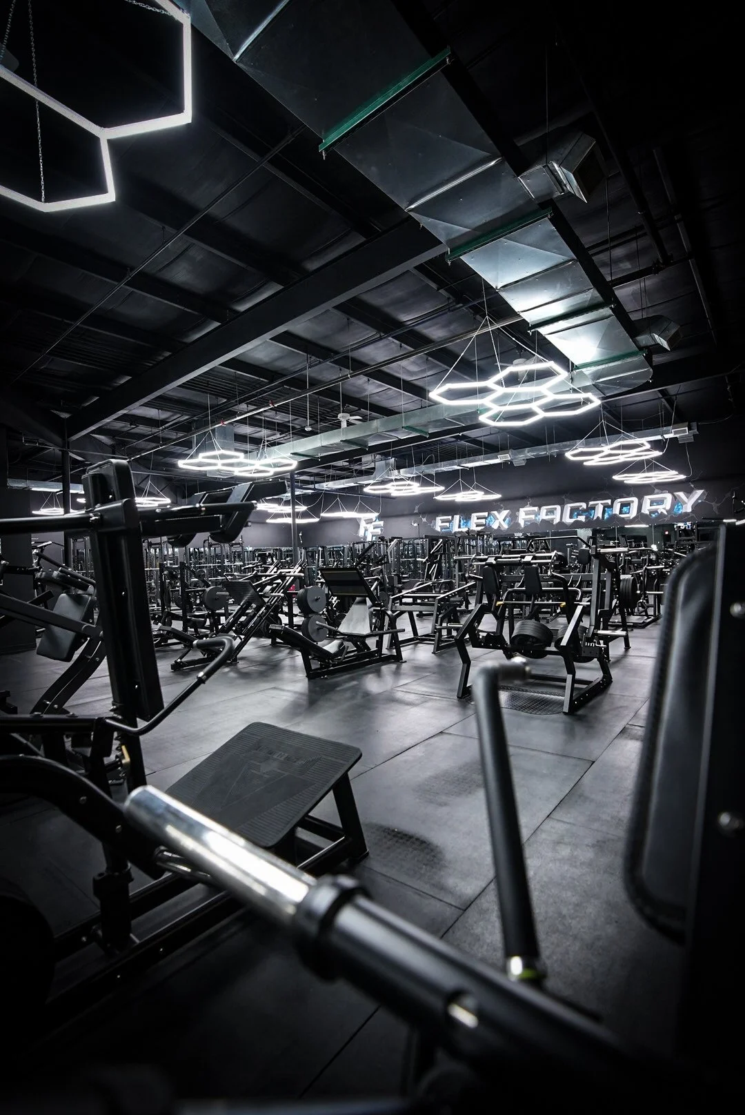 Interior of a modern, black-designed gym with various exercise equipment and illuminated hexagonal ceiling lights.