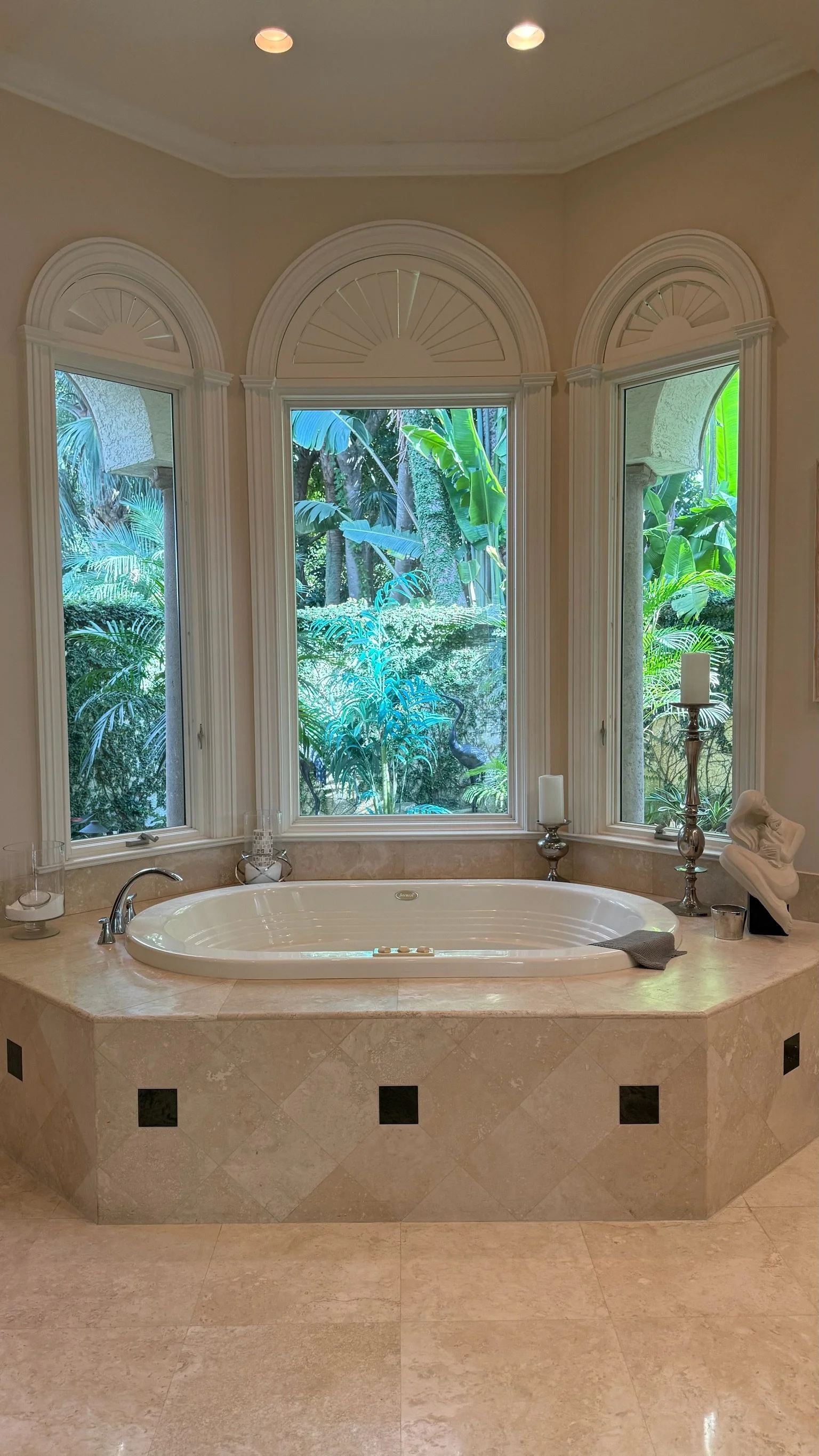 A luxurious bathroom with a built-in whirlpool bathtub surrounded by beige marble tiles, large bay window with lush green foliage outside, and decorative candles and sculptures on the ledge.