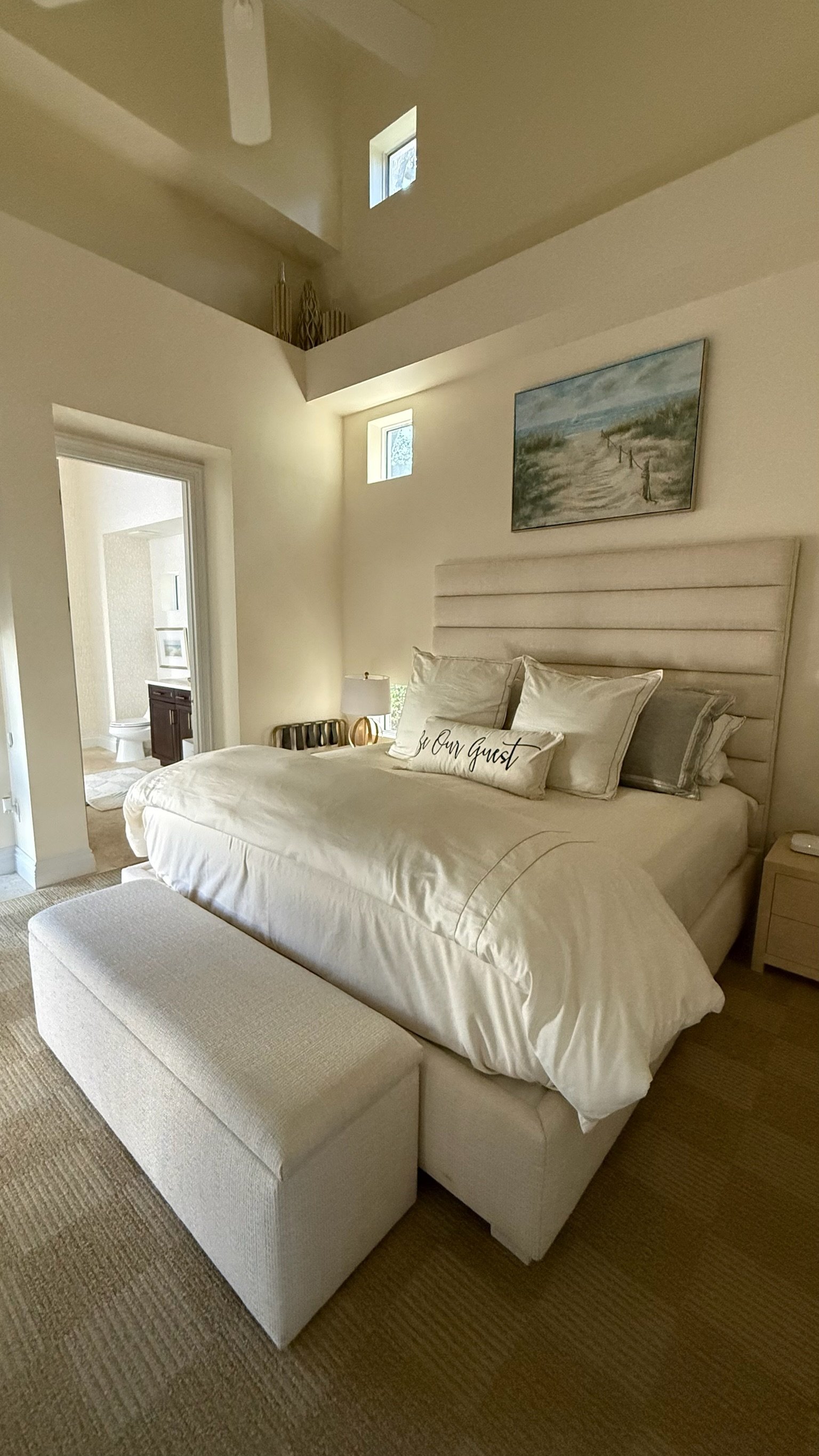 A cozy bedroom with a large bed, beige upholstered headboard, and a bench at the foot of the bed. The bed has white bedding and pillows, including one decorative pillow with