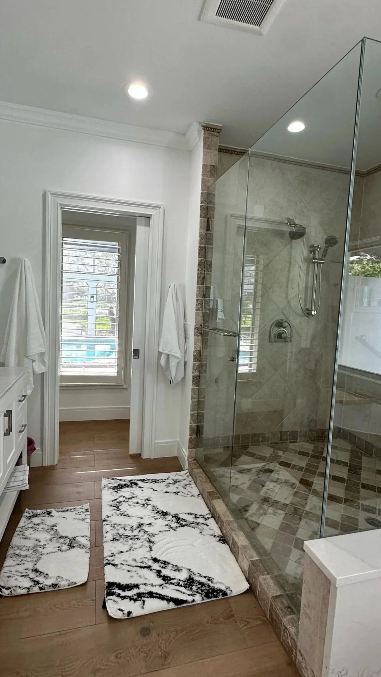 Modern bathroom with a glass shower, wooden flooring, white walls, and two patterned rugs outside the shower.