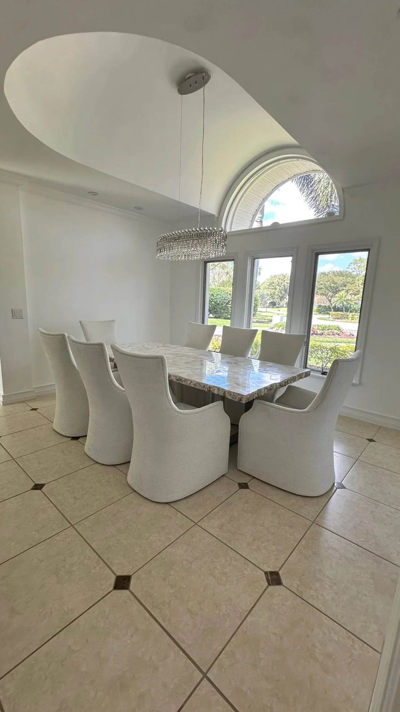 A bright dining room with a large marble table surrounded by eight white upholstered chairs. There is a crystal chandelier hanging above the table and a large arched window bringing in natural light, showing greenery outside.