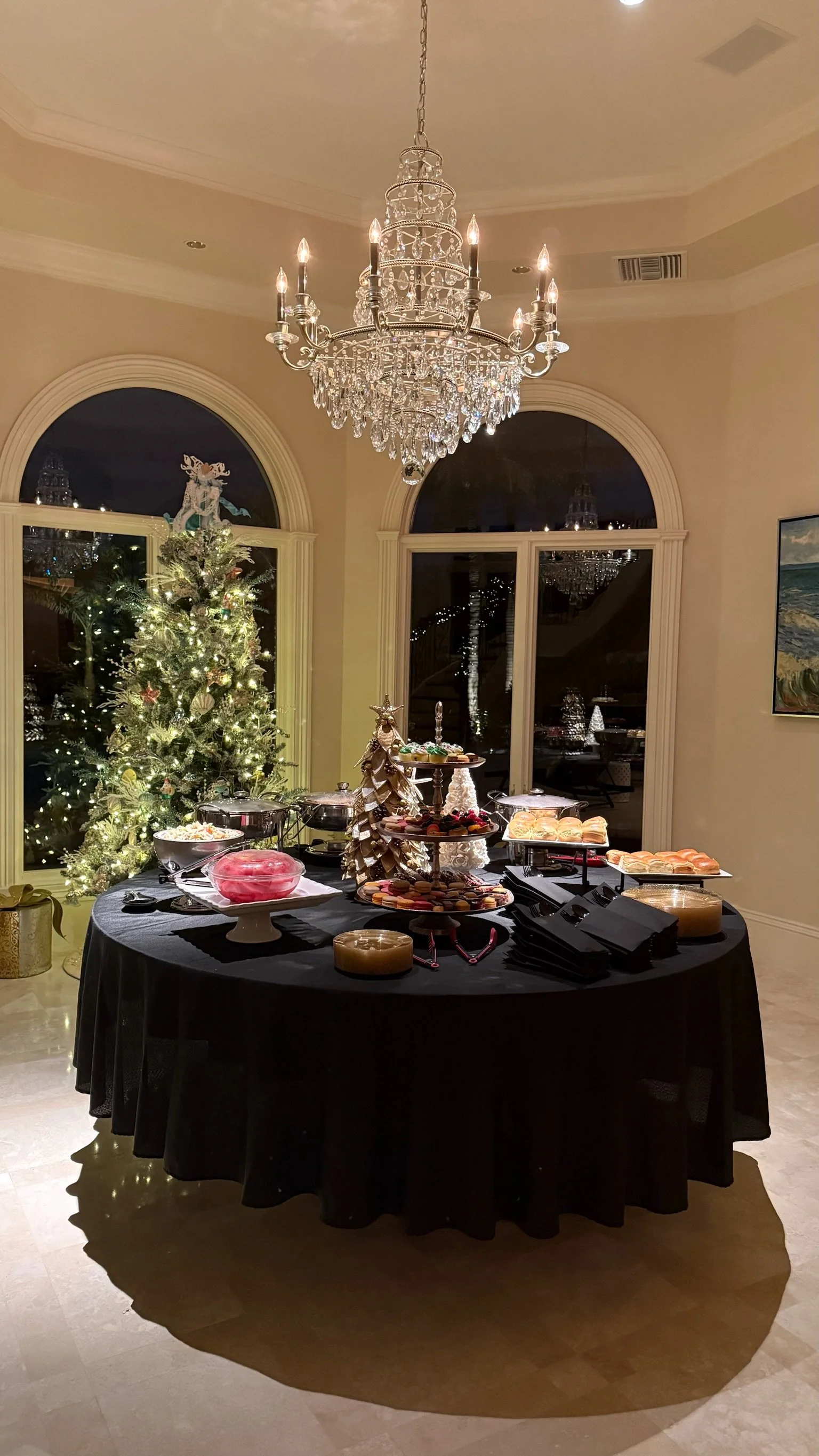 A Christmas party setup with a decorated Christmas tree and a table of desserts in a luxurious room with chandelier lighting.