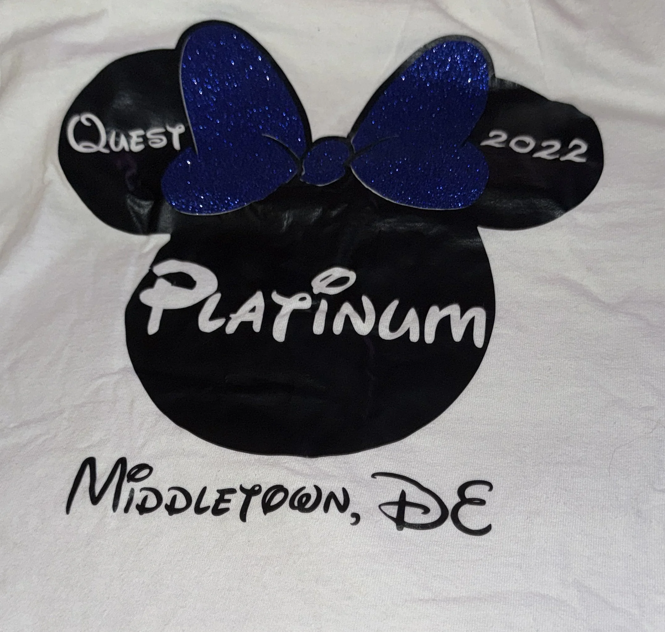 A black circular badge with a blue glittery butterfly on top, surrounded by black Mickey Mouse ears. The badge has the words 'Quest 2022' on the ears and 'PLATINUM' in the center. Below the badge, the words 'Middleton, DE' are written on a white surface.