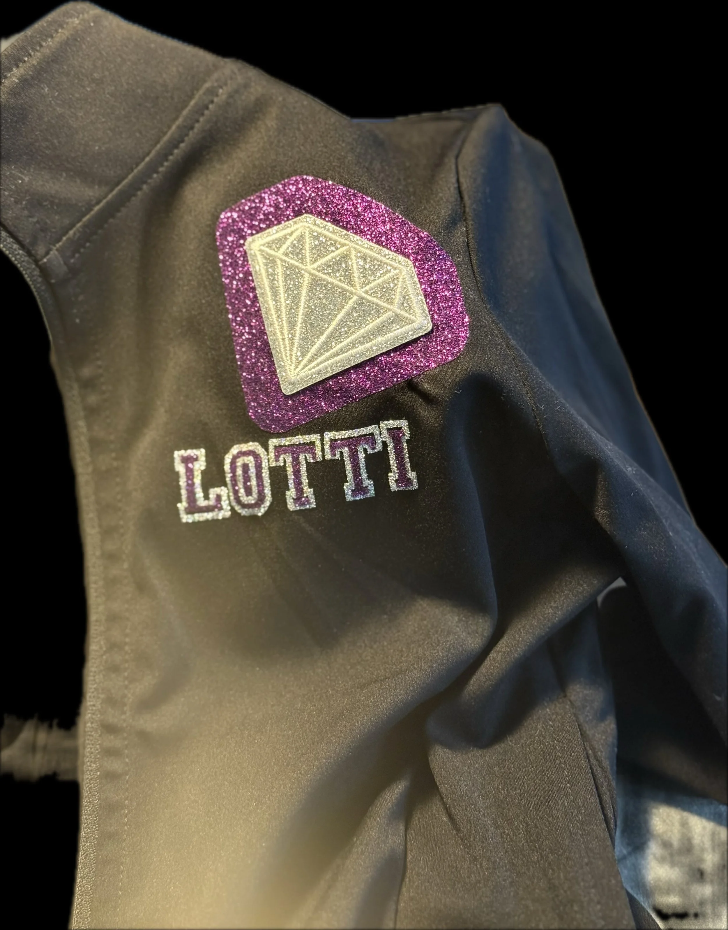 Close-up of a black jacket with a pink glittery diamond patch and the word 'LOTTI' in purple and white.