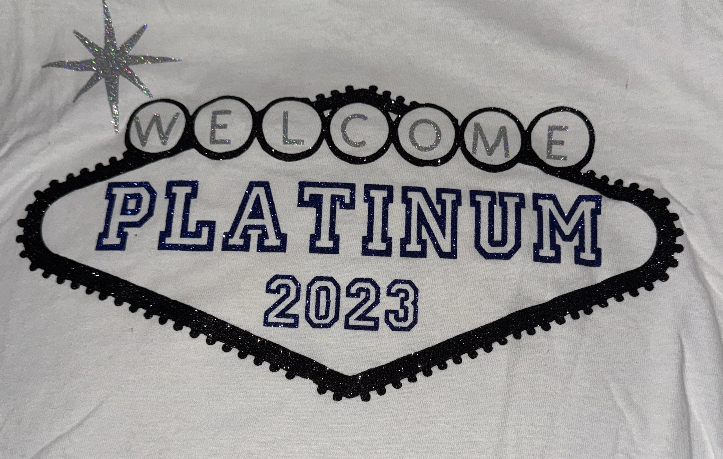 A white T-shirt with embroidered text and graphics reading 'Welcome Platinum 2023,' decorated with a glittery star and a black-bordered badge design.