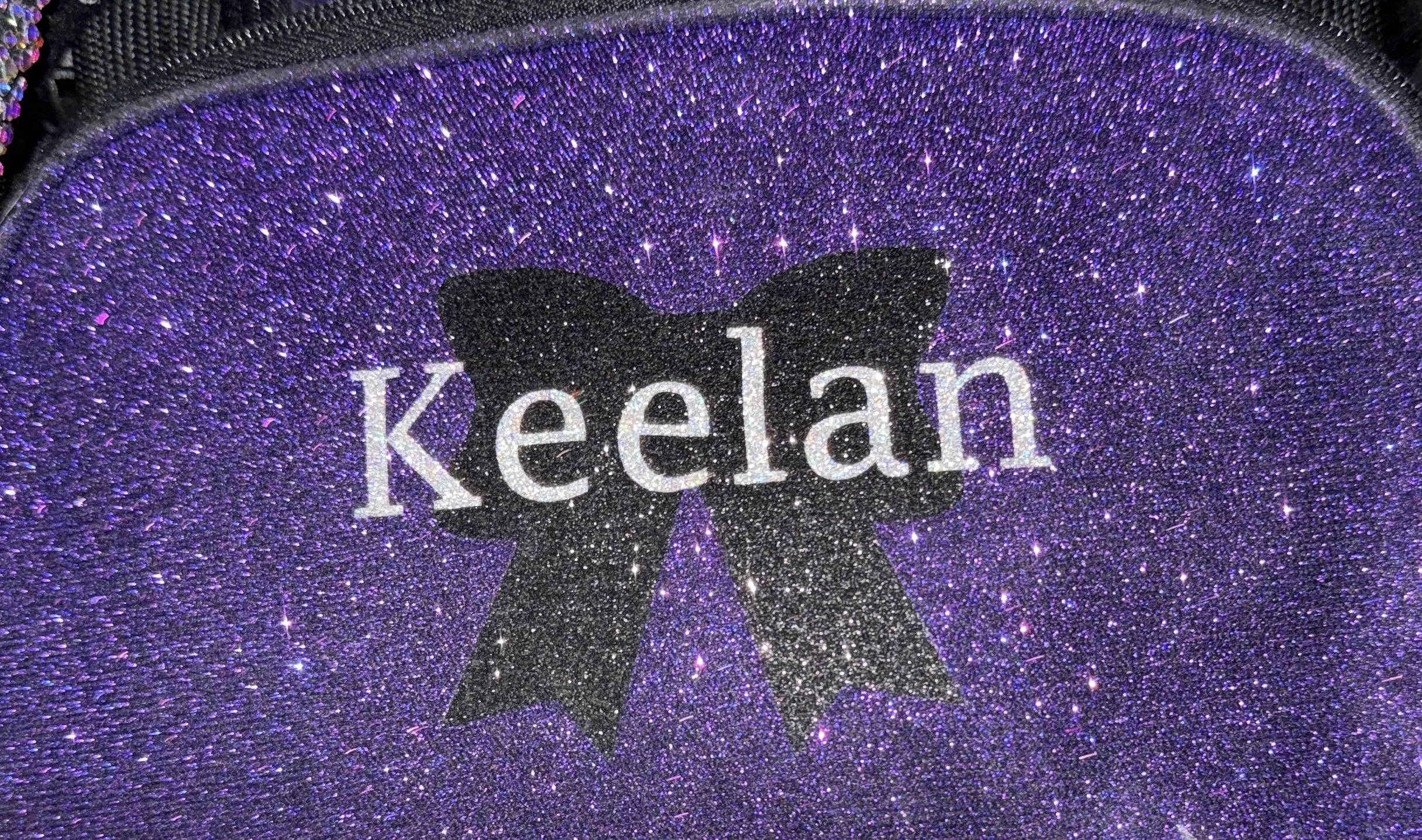 Purple glittery surface with a black bow and the name 'Keelan' written across it.