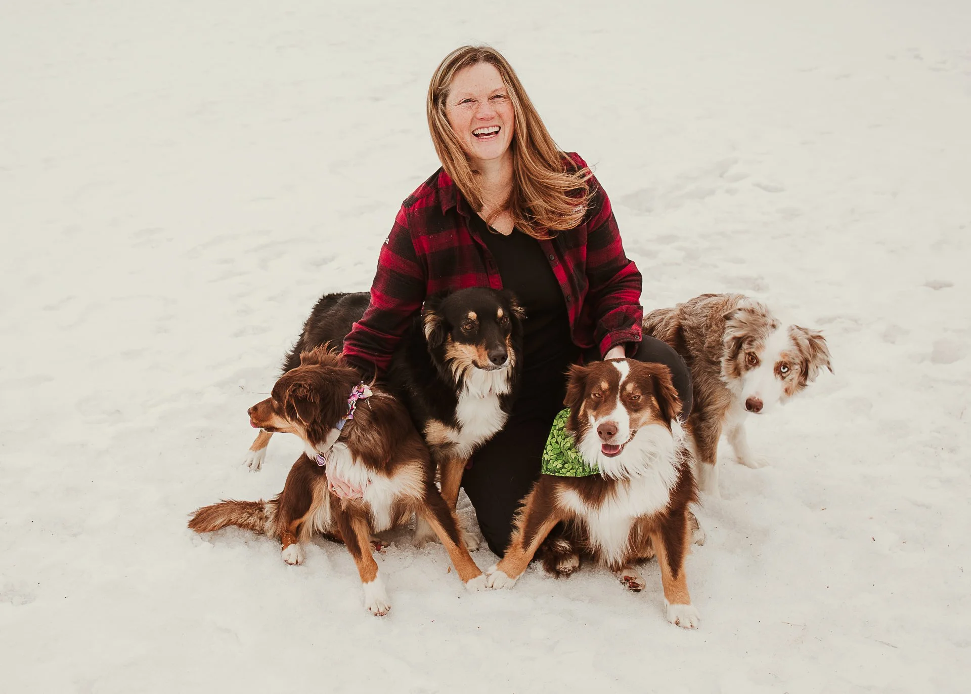 A woman with short blonde hair, wearing a sleeveless orange top and beige pants, sitting on a beige bench, writing in a notebook. There is a large green plant and a framed abstract art piece with earthy tones behind her, and sunlight casting shadows on the white wall.