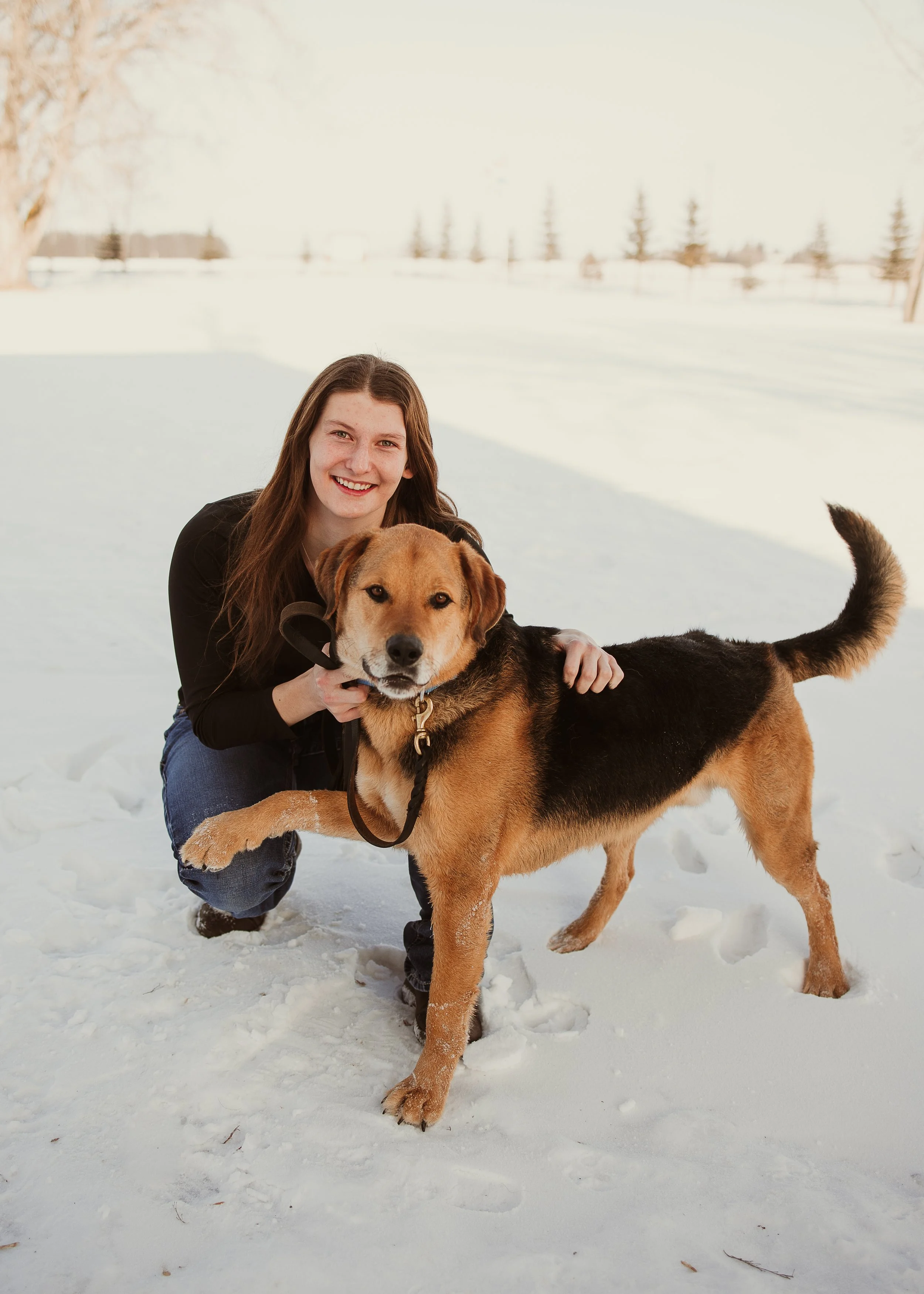 A woman with long brown hair, dressed in black, kneeling in the snow, smiling, holding a large black and brown mixed-breed dog with a leash in a snowy outdoor setting with trees and open space.