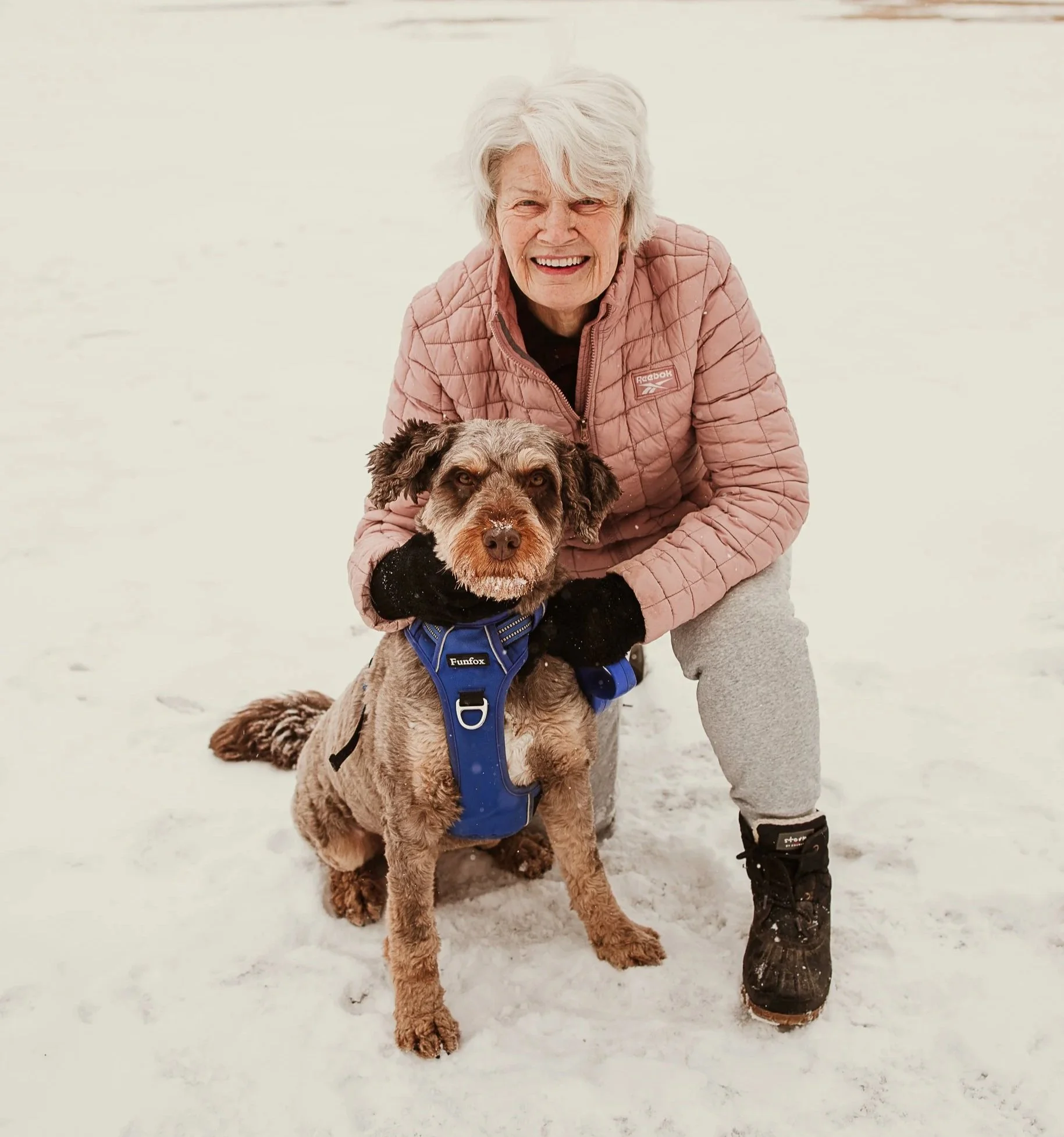 A man with gray hair wearing a blue short-sleeve sweater and beige pants sitting on a beige bench against a plain wall, with shadows cast on the wall.