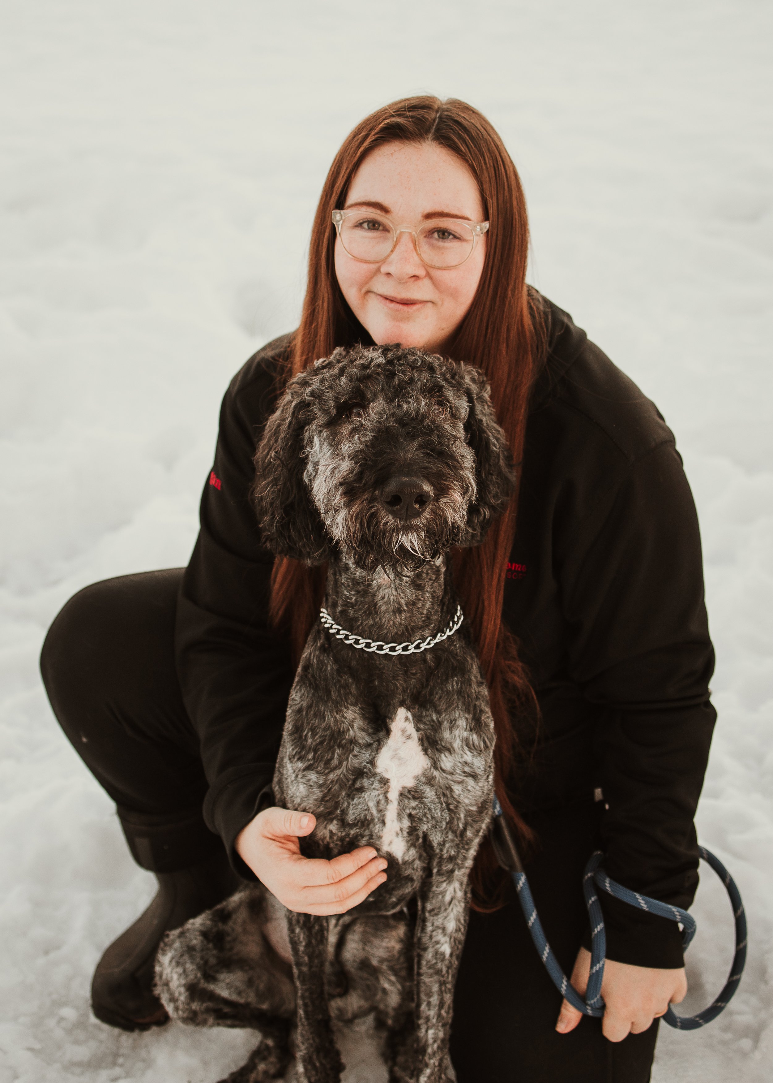 A young woman with long red hair, glasses, and a black hoodie standing on snow with a black and white dog on a leash outdoors.