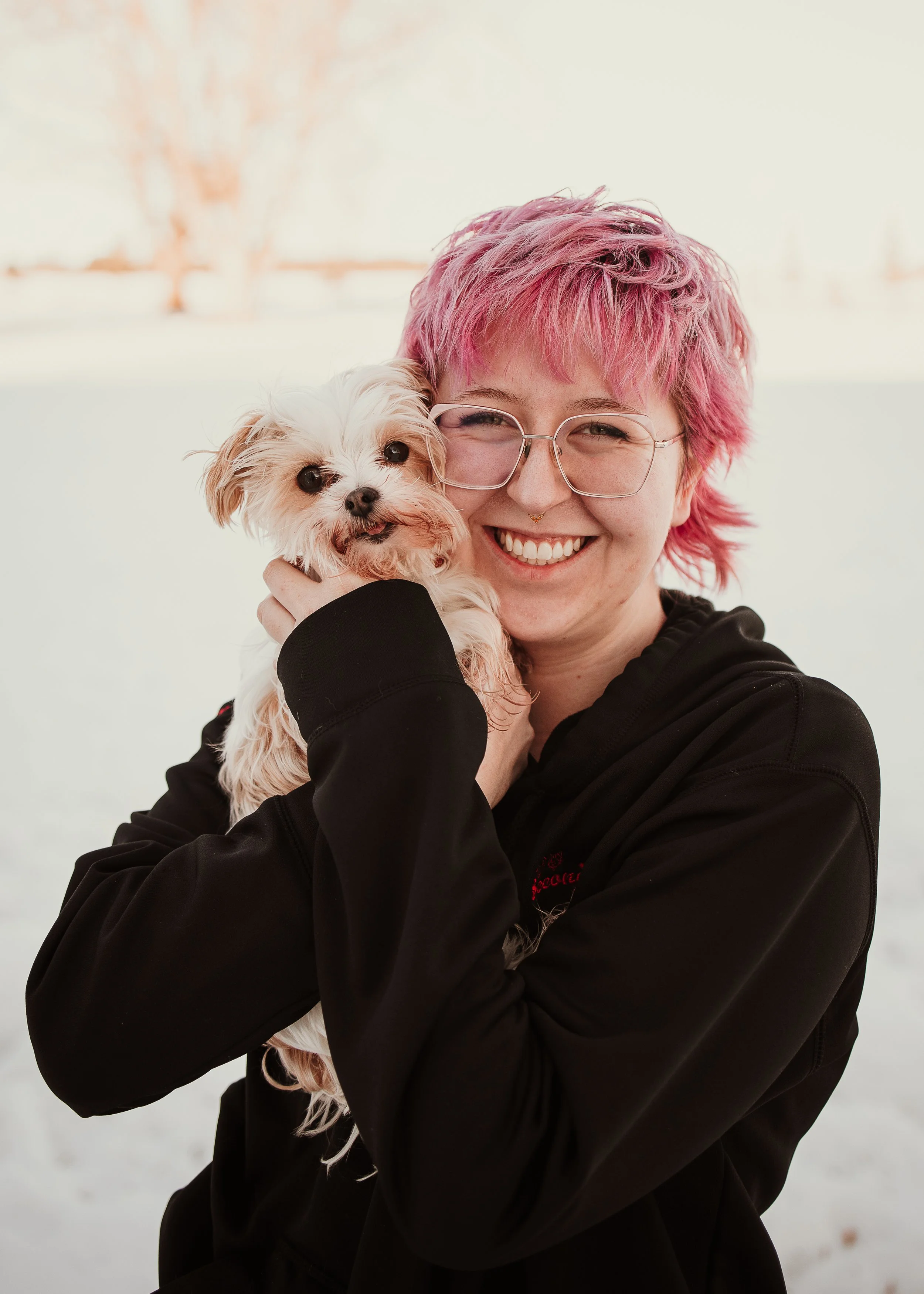 Person with pink hair, glasses, and a black hoodie holding a small fluffy dog outside in snow, smiling