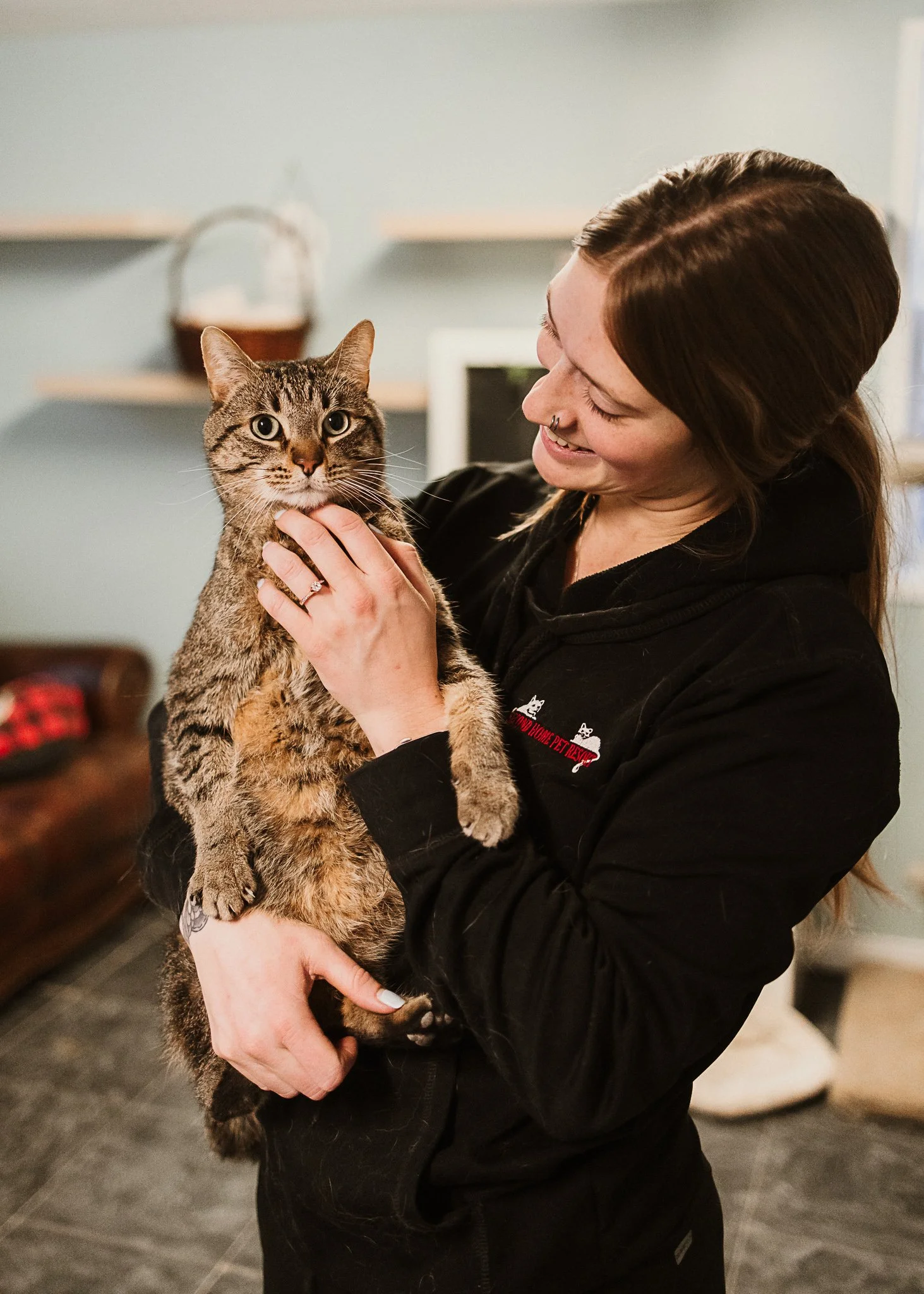 A female Second Home Pet Resort staff member holding a tabby cat indoors, smiling and looking at the cat