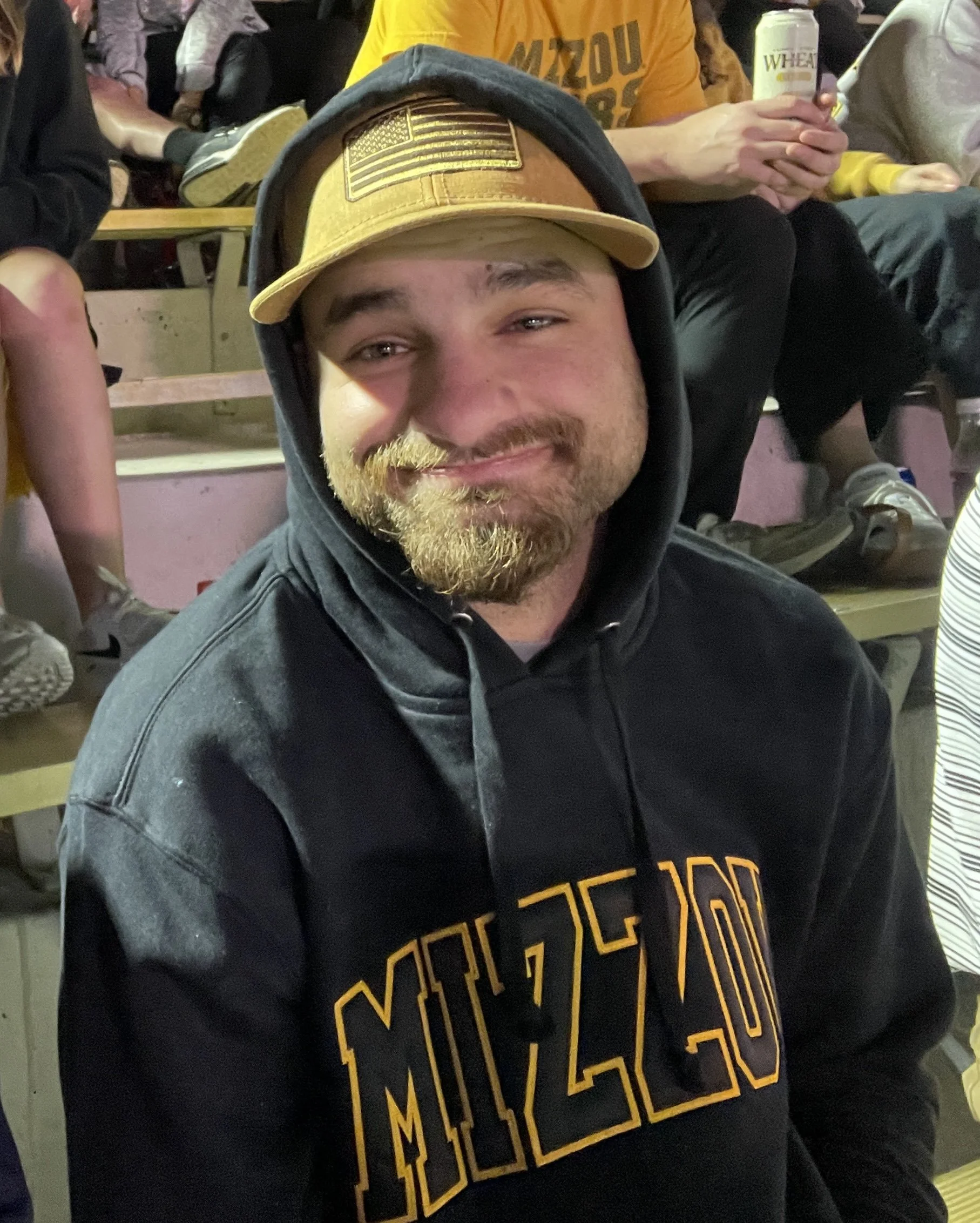 A young man with a beard and mustache wearing a black hoodie with MIZZOU written in yellow, and a tan baseball cap with an American flag patch, sitting in a crowd at an event.