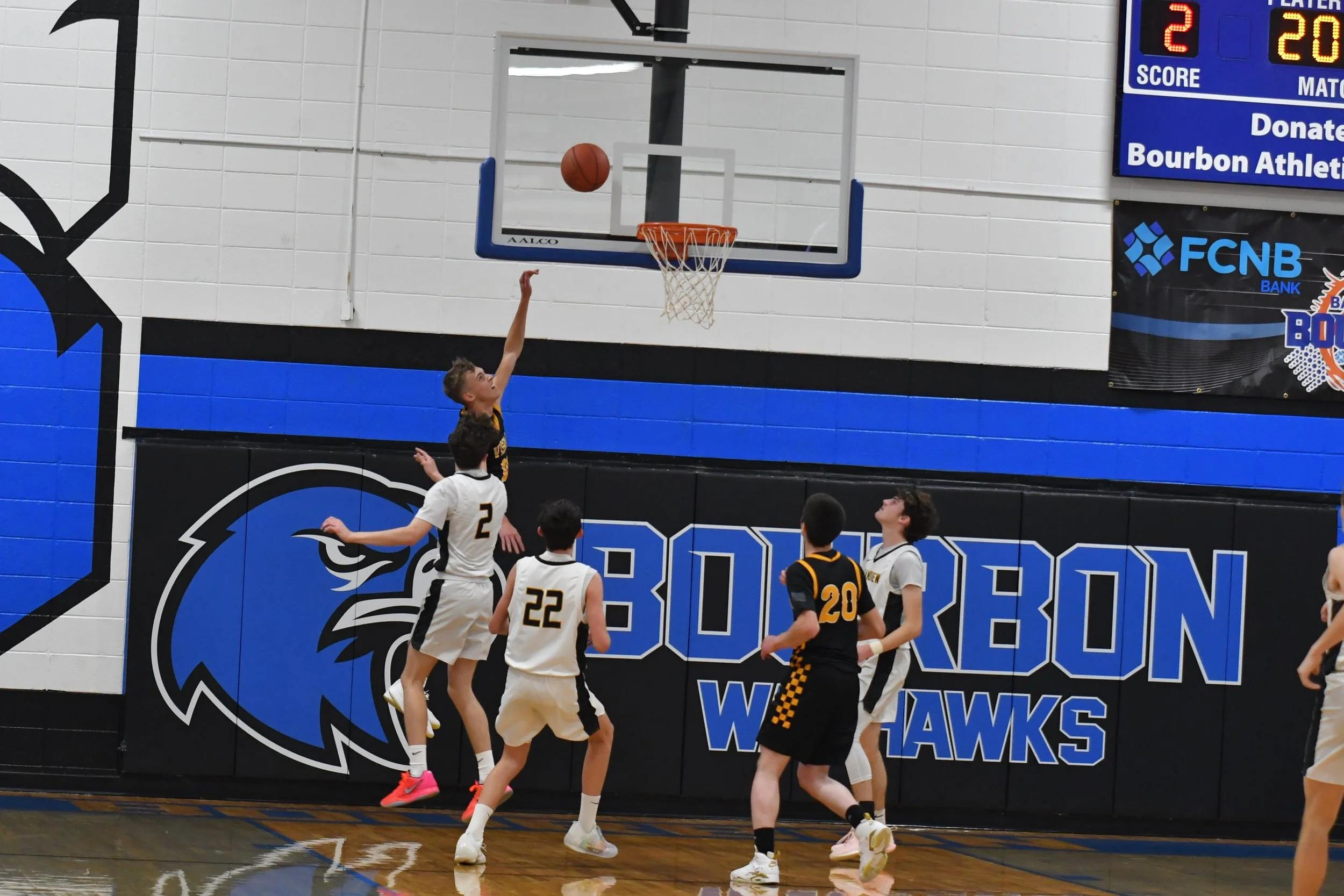 A basketball game in progress with a player in black, number 20, jumping towards the basket for a shot while five players in white and black uniforms watch or attempt to block. The gym has blue and black coloring with a logo that says 'Bourbon Wolver