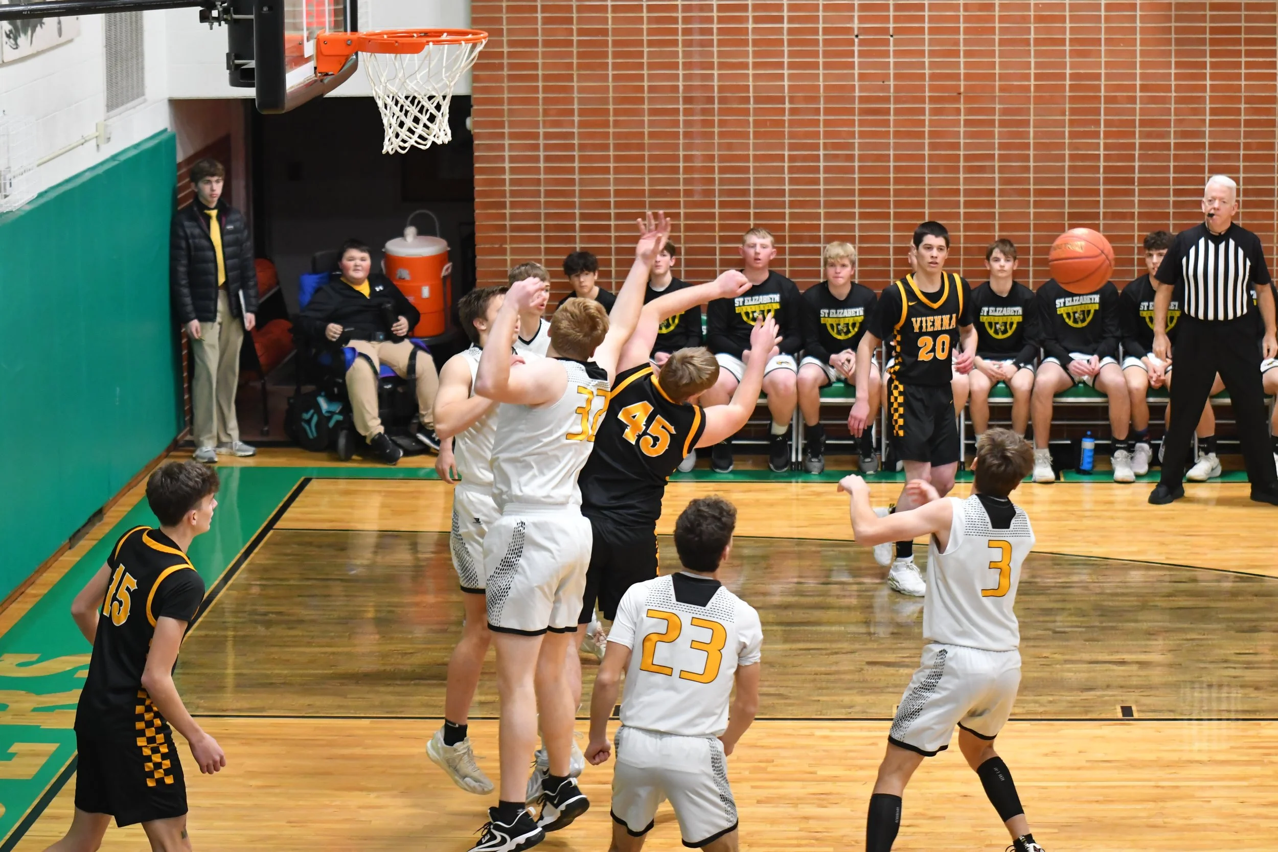 A basketball game in progress with players jumping to reach the ball near the basket, while players and a referee watch. Spectators and team members sit on the bench in the background.