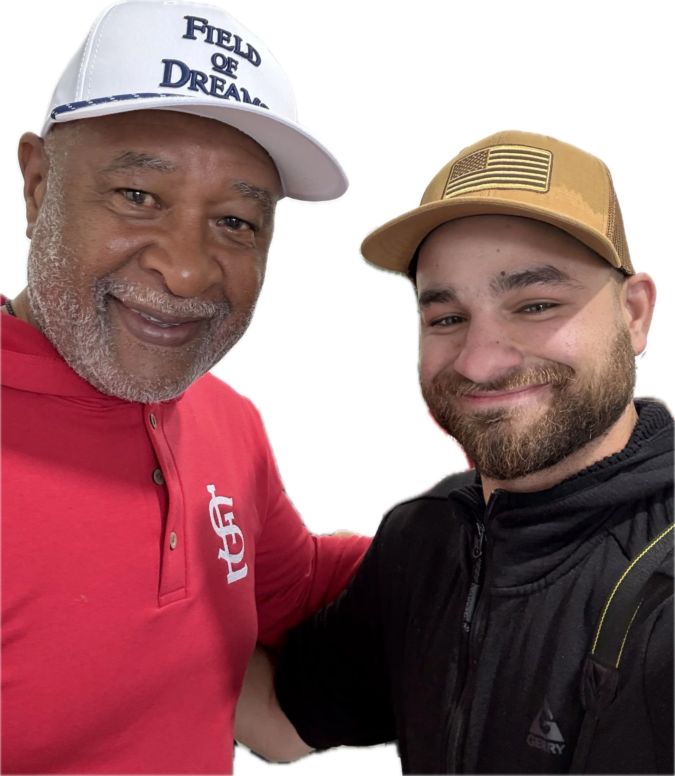 Two smiling men taking a selfie. The man on the left has gray hair and beard, wearing a white cap with blue text and a red shirt. The man on the right has a beard and is wearing a tan cap with a U.S. flag patch and a black jacket.