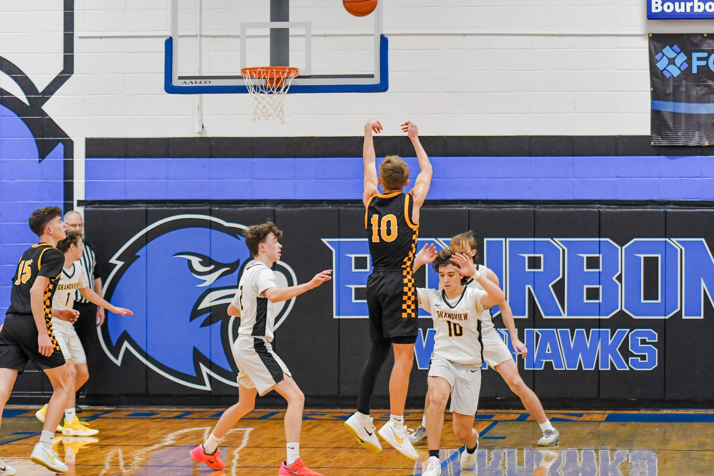 Young basketball players during a game in an indoor gym with a "Burbank Wildcats" banner and logo. Player wearing number 10 is shooting, others are defending or preparing for rebound.
